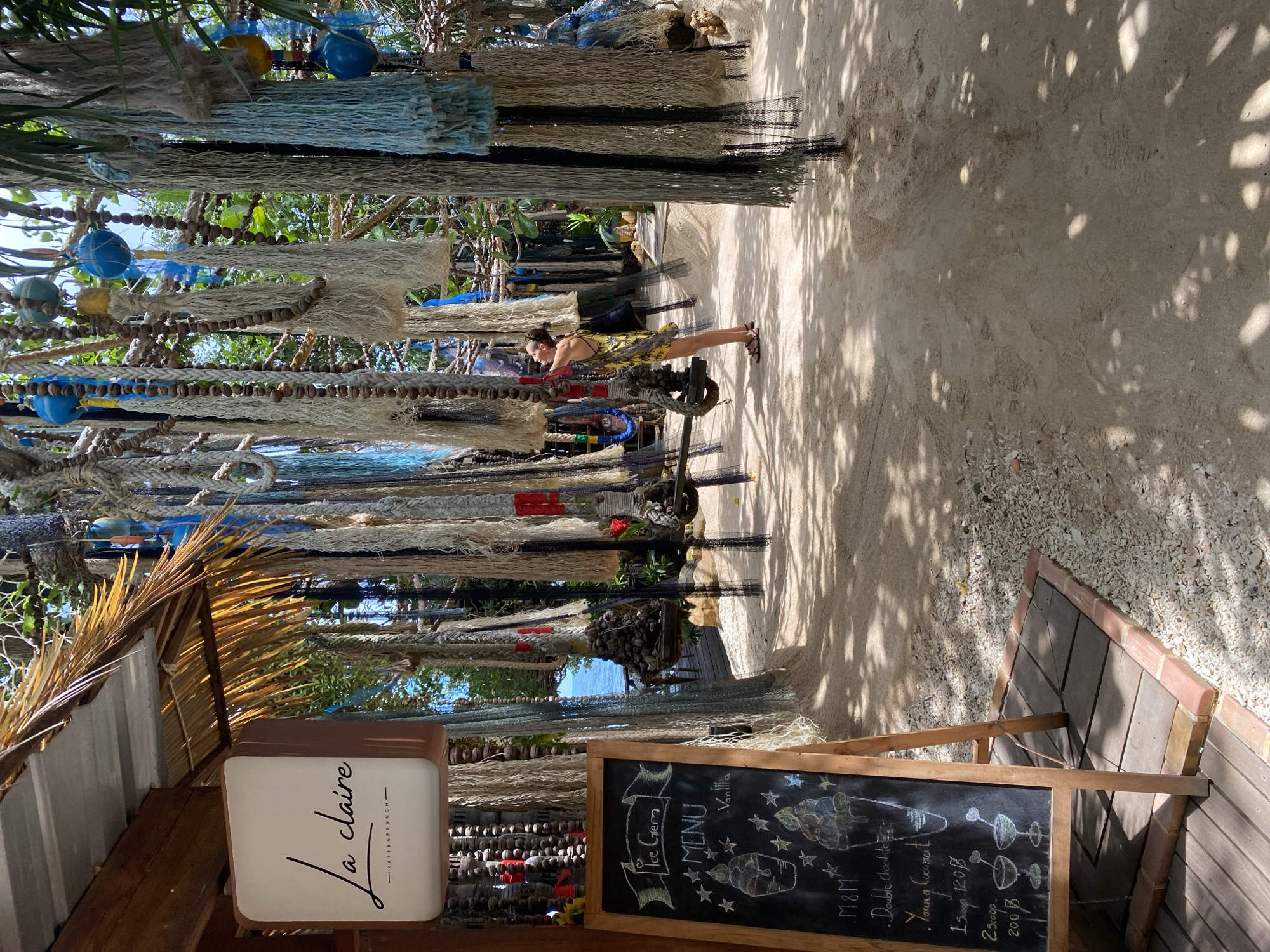 An outdoor, sandy area in front of a building features a woman in a yellow dress examining hanging decorations. The decorations are made of ropes, nets, and blue and white beads, strung from tree branches. To the left, a chalkboard menu displays ice cream offerings. The sign above the menu reads "La claire - KAFFESBRUNCH -". The background shows a glimpse of the ocean and foliage. The scene appears to be in Koh Phangan, Thailand.