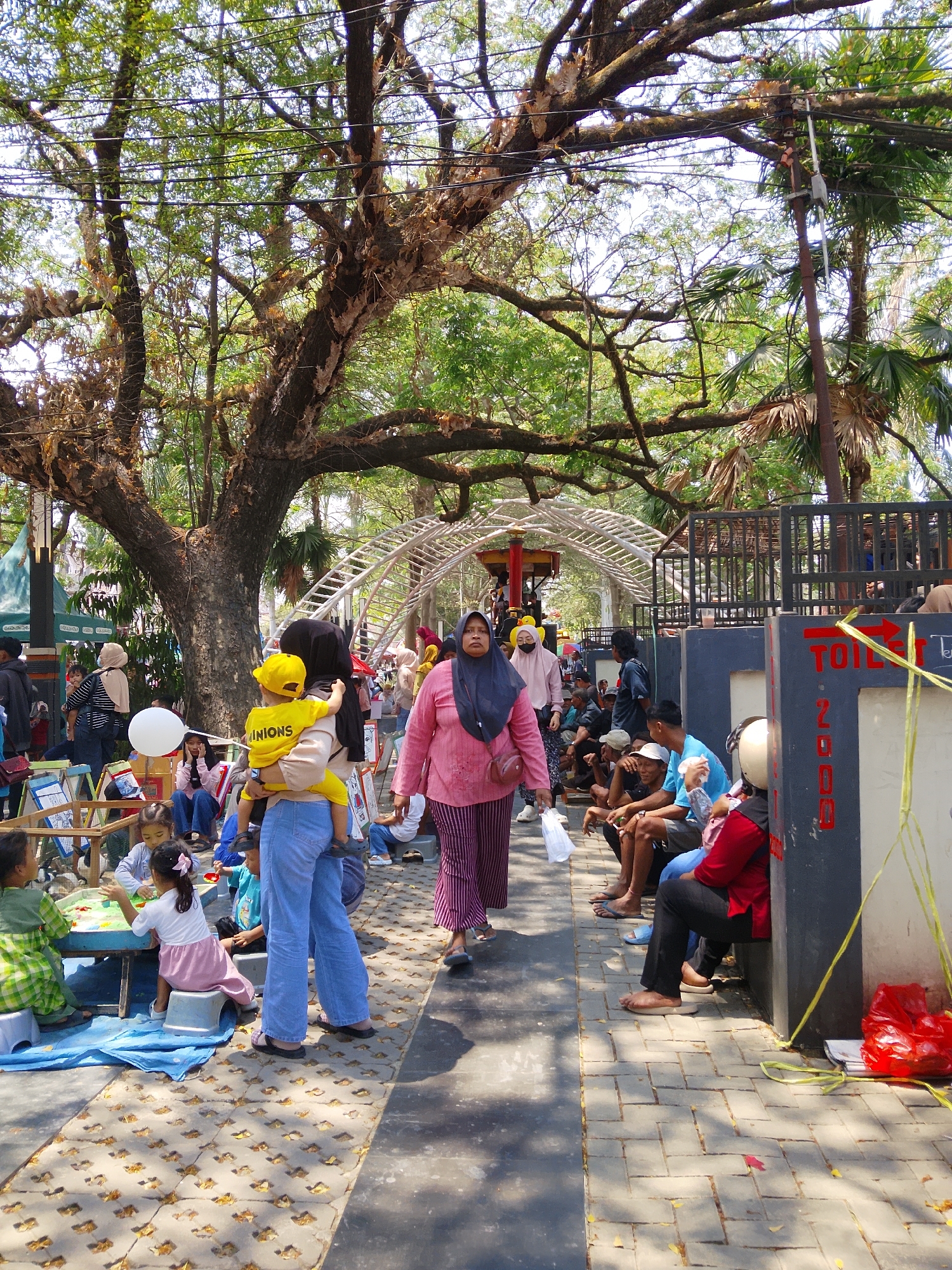 In Balun, Indonesia, a public outdoor space is filled with people under the shade of large trees. In the foreground, a woman in a pink hijab and a pink and white striped outfit walks towards the right of the frame, carrying a white plastic bag. To her left, a woman wearing a black hijab holds a young child wearing a yellow shirt and hat. The child's shirt has the word "MINIONS" printed on it. Several other people, including children, are seated or standing around tables, some engaging in activities. In the background, a white arch structure extends across the path, and a black metal fence encloses an area. A dark gray structure with "TOILETS" written on it is visible on the right side of the image, with people sitting near it. The ground is paved with interlocking bricks, and fallen leaves are scattered on the surface. Sunlight filters through the tree canopy, creating dappled shadows.