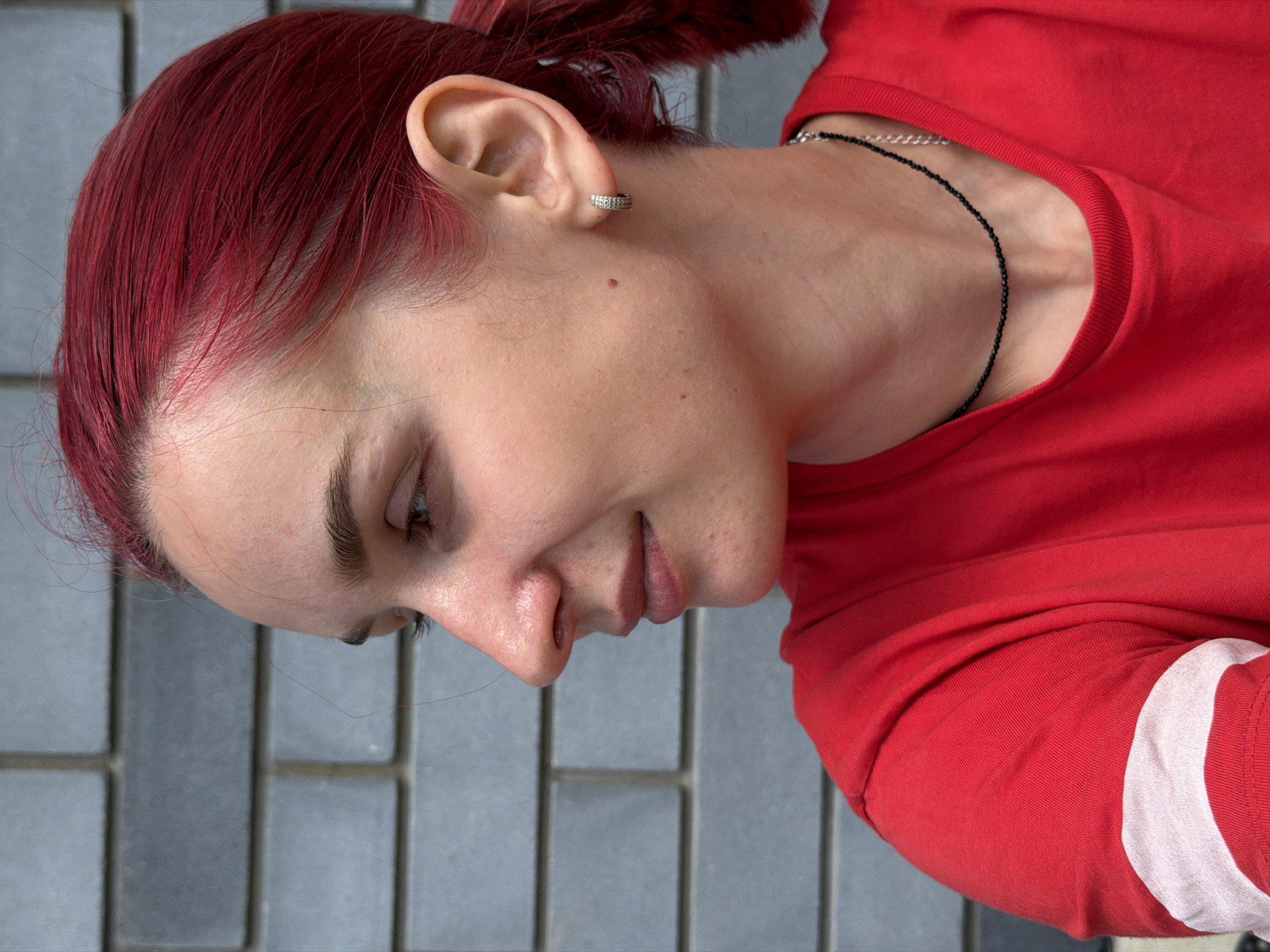 A young woman with bright red hair is shown in profile, looking to the left. She is wearing a red t-shirt with a white stripe on the sleeve. She has a small hoop earring in her left ear and a thin black necklace. The background is a gray brick wall. The lighting suggests it is daytime outdoors. The location is Chaikovsky, Russia.