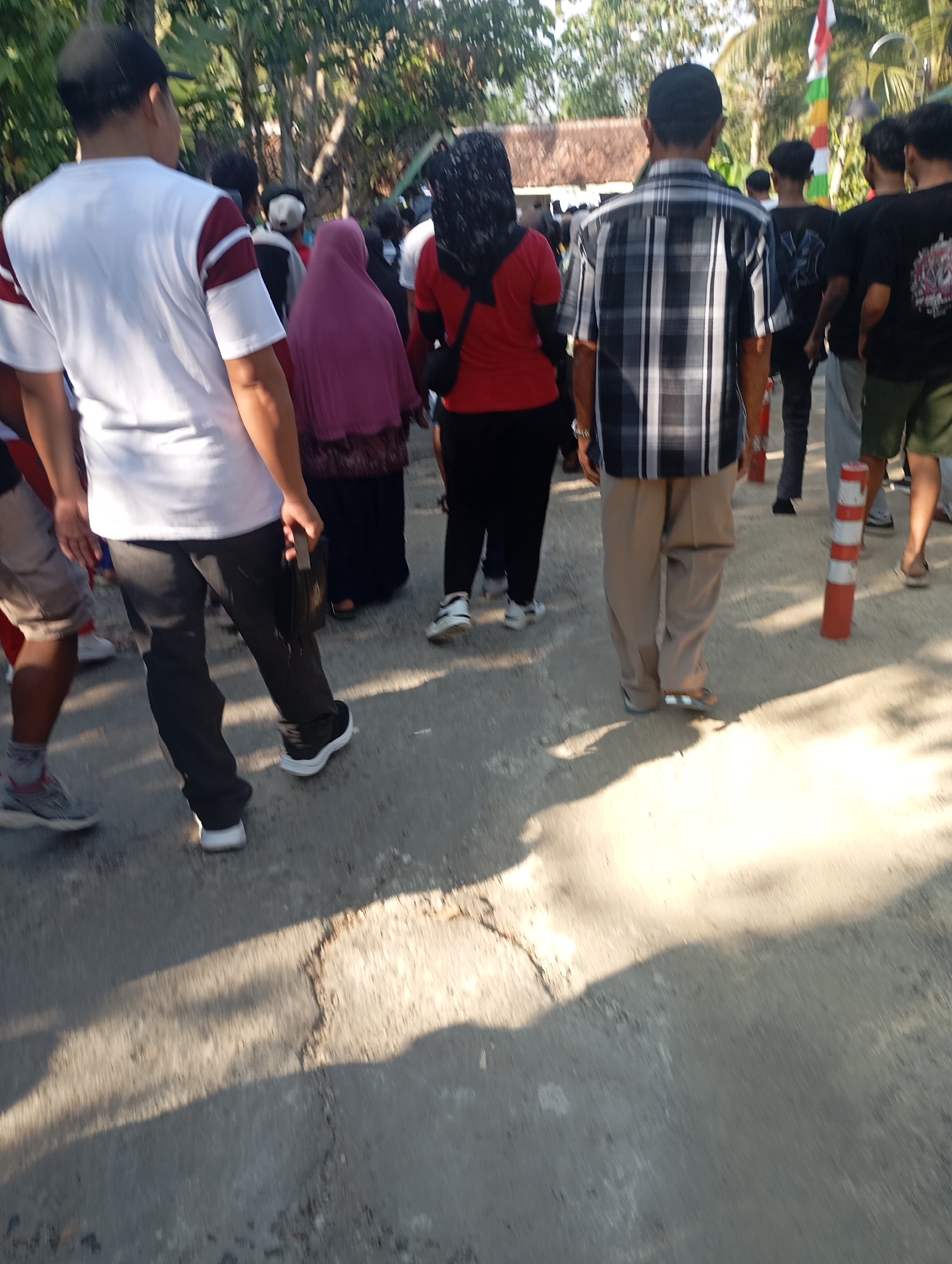 A group of people are walking on a street in Ketepung, Indonesia. The sun is shining, casting shadows on the ground. The people are walking in the same direction. A man in the foreground is wearing a white t-shirt with maroon stripes on the sleeves and black pants. He is holding a dark bag in his right hand. To his left, a person in a pink hijab is walking, followed by a person in a red shirt. To the right of the man in the foreground, a man in a black and white plaid shirt is walking. Behind them, several other people are visible, some wearing black t-shirts. In the background, trees and a building with a thatched roof can be seen. Several orange and white traffic cones are placed along the side of the street. The street appears to be paved with asphalt and is cracked in some areas. A flag with red and white colors is visible in the background.