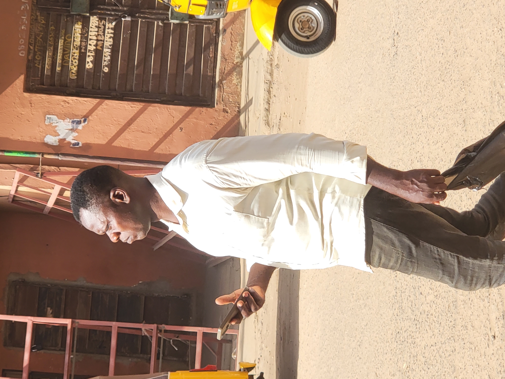A medium outdoor shot captures a man standing on a paved surface next to a building in what is presumed to be the city of Maiburiji, Nigeria. The man is facing left and looking down at the mobile phone he holds in his right hand. He is wearing a light-colored long-sleeved shirt and dark pants. The building is adjacent to the street, and a yellow motorcycle or motorized vehicle is parked alongside it. A metal grid structure is also visible behind the man. The ground appears to be dry and dusty.