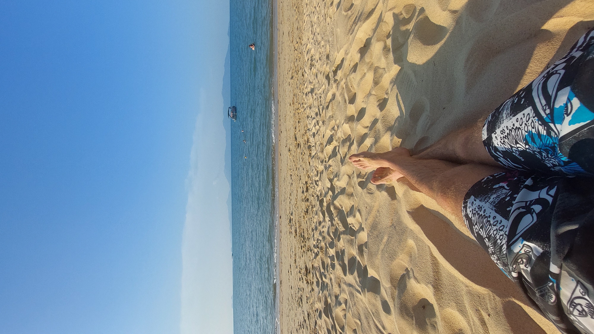 This image captures a serene beach scene from a first-person perspective, with the viewer's legs and feet prominently in the foreground. The person is relaxing on golden, finely textured sand, wearing black, white, and blue patterned swim trunks. Sharp shadows stretch across the sand, indicating bright, direct sunlight, likely during the late morning or afternoon.
The sandy beach extends towards the horizon, meeting a calm, clear blue-green sea. Gentle waves can be seen breaking softly at the shore. In the water, a person's head is visible, swimming, and further out, a small boat is anchored. Several buoys are scattered across the water's surface. Distant land or mountains are faintly visible along the horizon line, blurring into the bright blue sky above. The sky is largely clear, with just a hint of very light cloudiness towards the right edge.
The scene conveys a sense of peaceful relaxation and leisure, typical of a vacation setting. Given the context, this tranquil beach is located in Baan Mae Nam, Thailand. No visible text is present in the image.