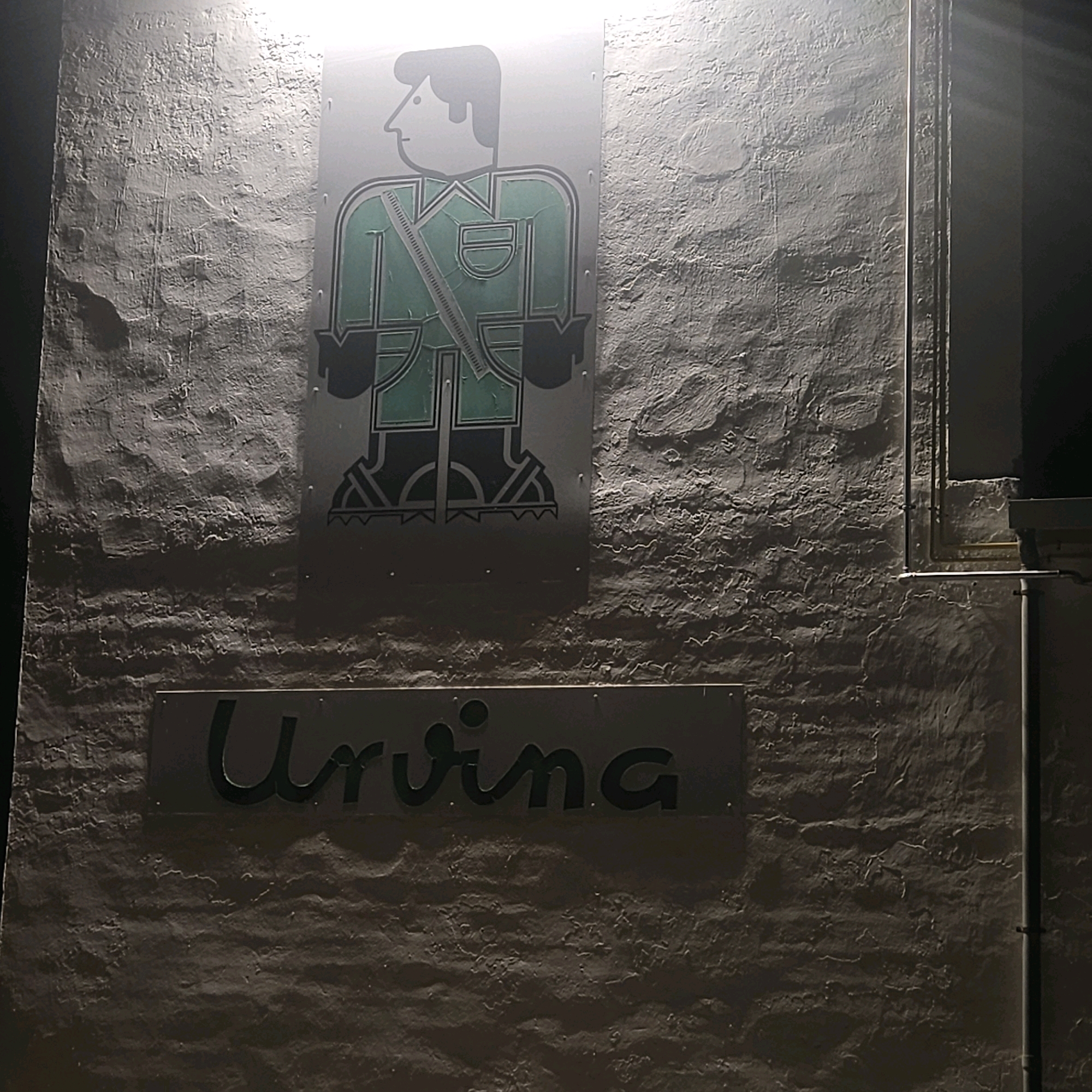 In Polígono Industrial Los Leones, Spain, a rough, textured wall is illuminated by light. Affixed to the wall is a metallic sign depicting a stylized figure wearing a green uniform with a diagonal strap across its chest. Below this figure, another metallic sign displays the name "Urvina" in stylized black lettering. The overall scene suggests an exterior signage on a building within an industrial park.
