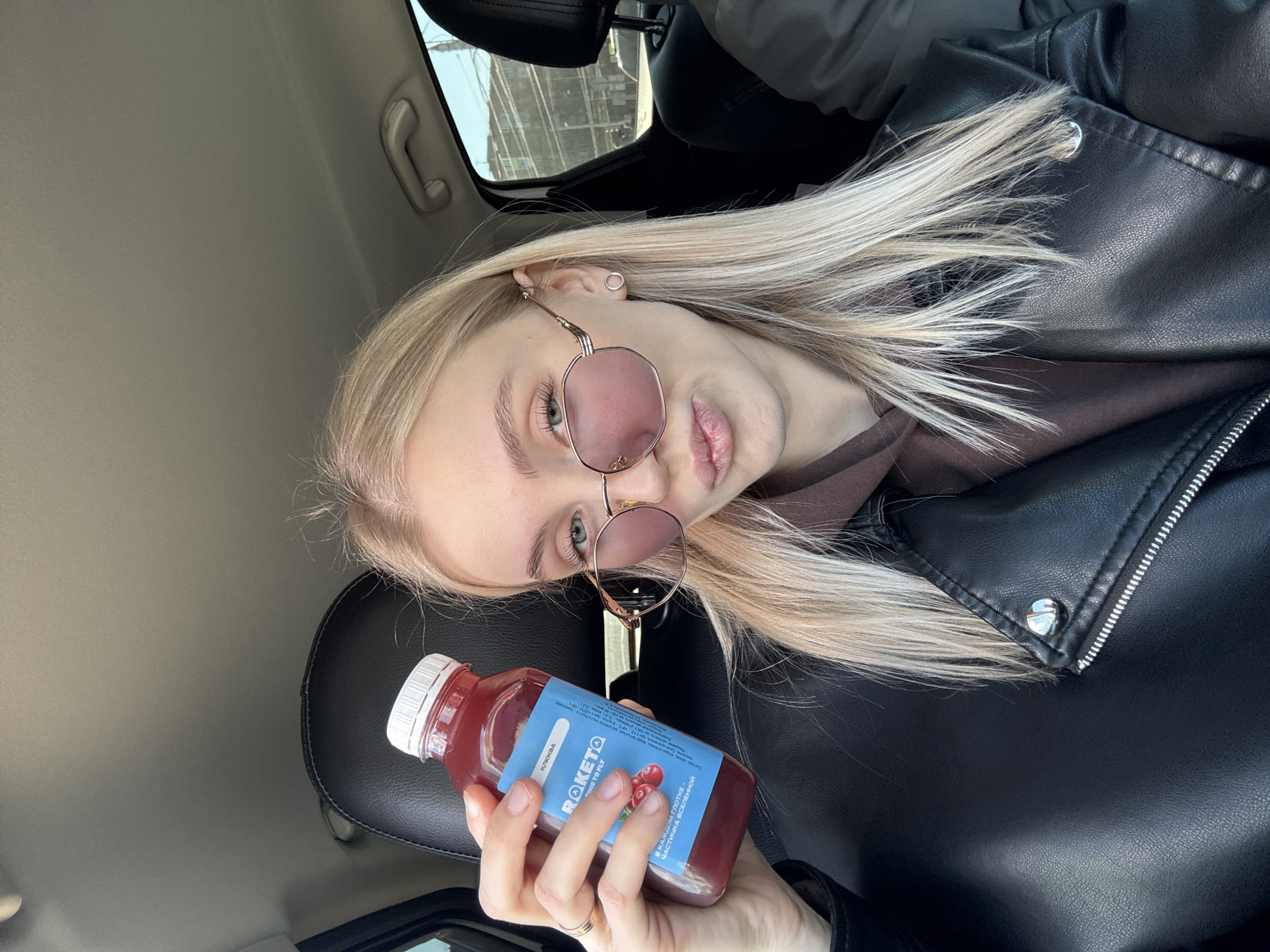 A young, blonde-haired woman is positioned in the driver's seat of a car, holding a bottle of "ROKETO" cranberry drink. The bottle has Russian text. She wears round sunglasses with pink-tinted lenses and is dressed in a brown sweatshirt under a black leather jacket. The interior of the car is visible, including the headrest and ceiling. The image appears to be taken from inside the car, with the woman holding the drink up for the photo. There is no visible location confirming that this image was taken in the city of Izhevsk, Russia.