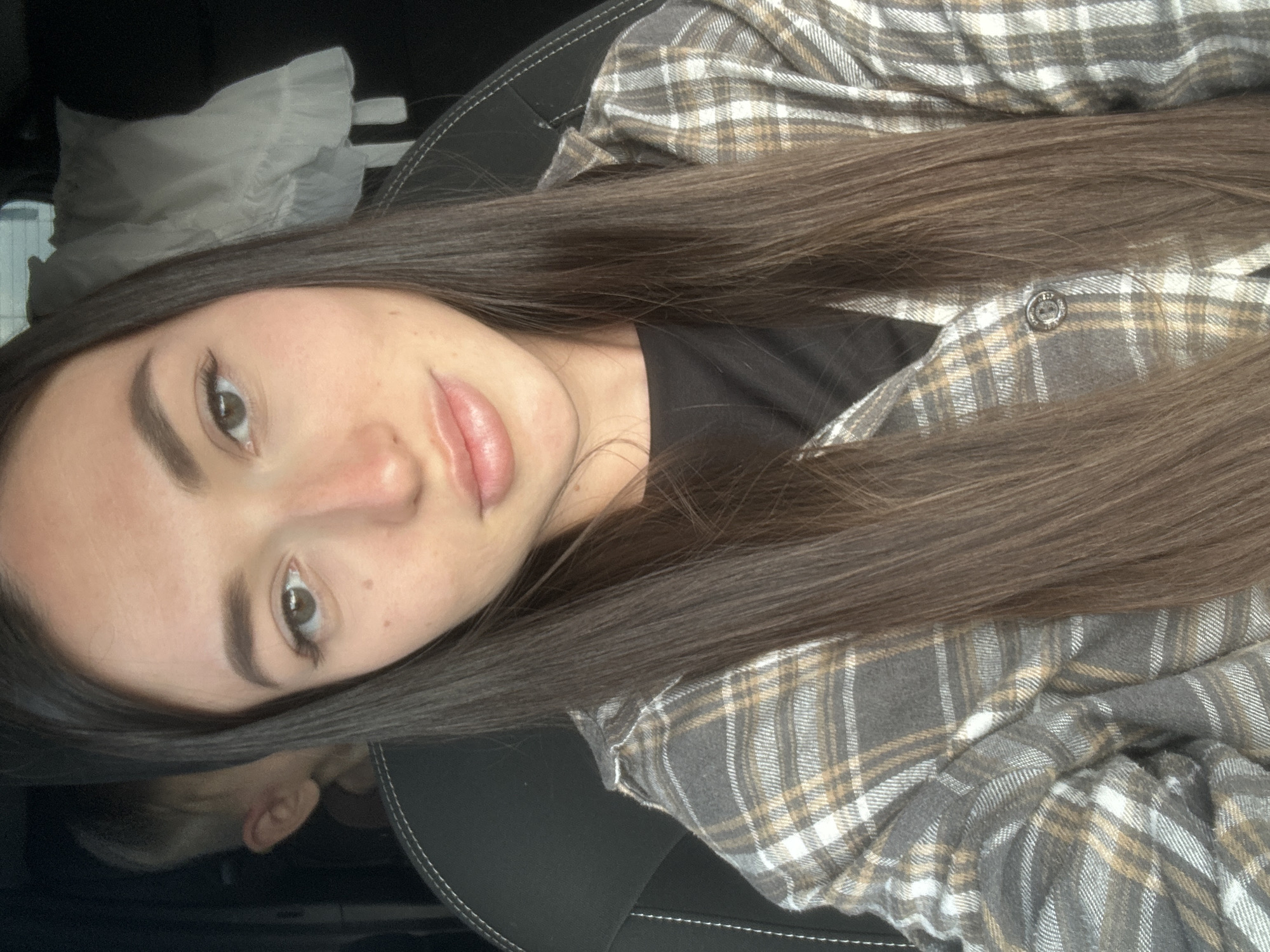 This is a selfie of a young woman with long, dark brown hair, wearing a grey and tan plaid shirt over a black t-shirt. She is sitting in a vehicle, indicated by the seatbelt visible on the left and the car seat behind her. A young boy, with short light brown hair, is partially visible in the background behind her left shoulder. The interior of the vehicle appears to be a dark color. There is a white frilly object on the right side of the frame, possibly a car accessory or clothing. The overall lighting suggests it's daytime. There is no discernible information in the image to indicate the location is Kazan, Russia.