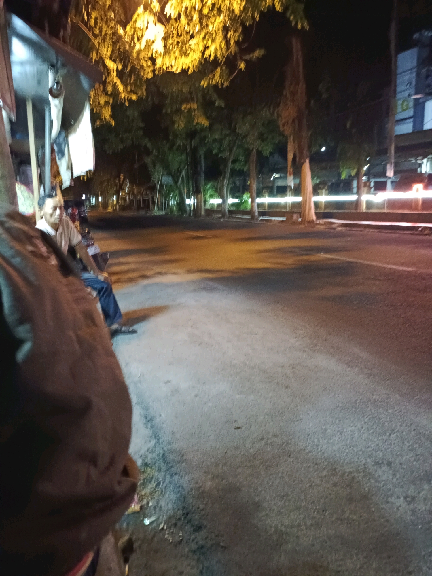 A medium shot is captured at night, focusing on a man seated on a motorcycle. The man is wearing a light-colored polo shirt and dark trousers. He is looking to his right. The motorcycle appears to be parked on the side of a street. Behind the motorcycle, a rudimentary awning-like structure is visible, with some fabric hanging from it. The street is paved and appears to be well-lit by artificial lights, casting shadows on the ground. Trees line the street, their leaves illuminated by the lights. In the background, there are buildings, some with illuminated windows. The overall scene suggests an urban environment at night, possibly a roadside stall or resting area. This image was taken in the city of Lamongan, Indonesia.