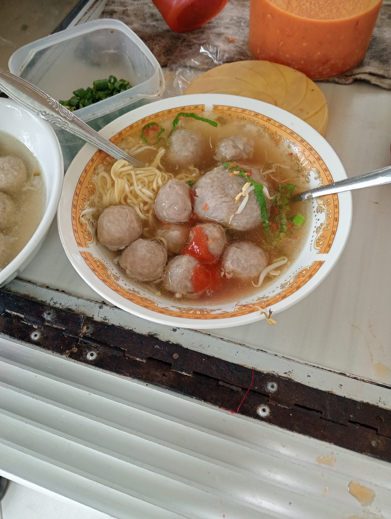 A close-up, overhead shot displays a bowl of Indonesian meatball soup, known as Bakso, in the city of Sidoharjo, Indonesia. The Bakso is served in a white bowl with an orange and gold floral pattern. The soup contains several large, round meatballs, a portion of noodles, and bean sprouts, garnished with chopped green onions and chili. A fork rests in the bowl, partially submerged. To the left of the main bowl, another white bowl holds a few meatballs in broth. In the background, a clear plastic container holds more chopped green onions, and a large orange, semi-spherical object, possibly a food item or container, is visible. The setting appears to be an outdoor food stall or a casual eatery, with a metallic surface and rusty hinges in the foreground, suggestive of a weathered, functional preparation area.
