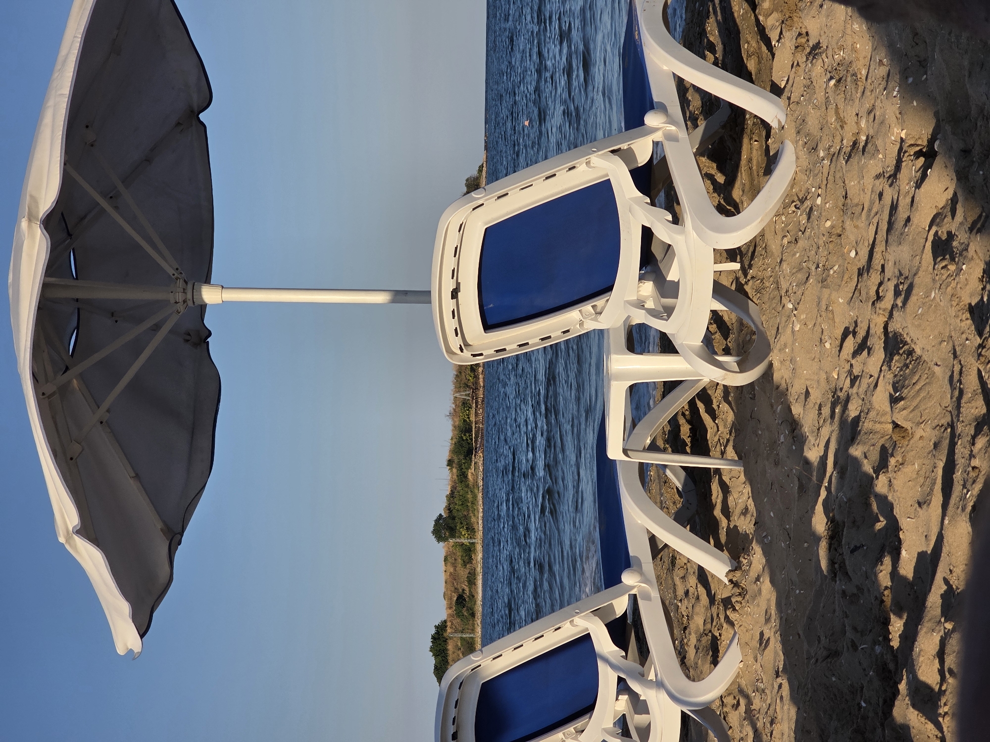 The image captures a serene beach scene at Chernomorets, Bulgaria. Two white plastic lounge chairs with blue cushions are positioned on the sand, facing the calm, blue sea. A large white umbrella provides shade over the chairs. The background features a shoreline with sparse vegetation and a clear blue sky. The sunlight casts long shadows across the sand, indicating it is either early morning or late afternoon. There are no people visible in the frame. The environment suggests a peaceful, unoccupied beach setting.