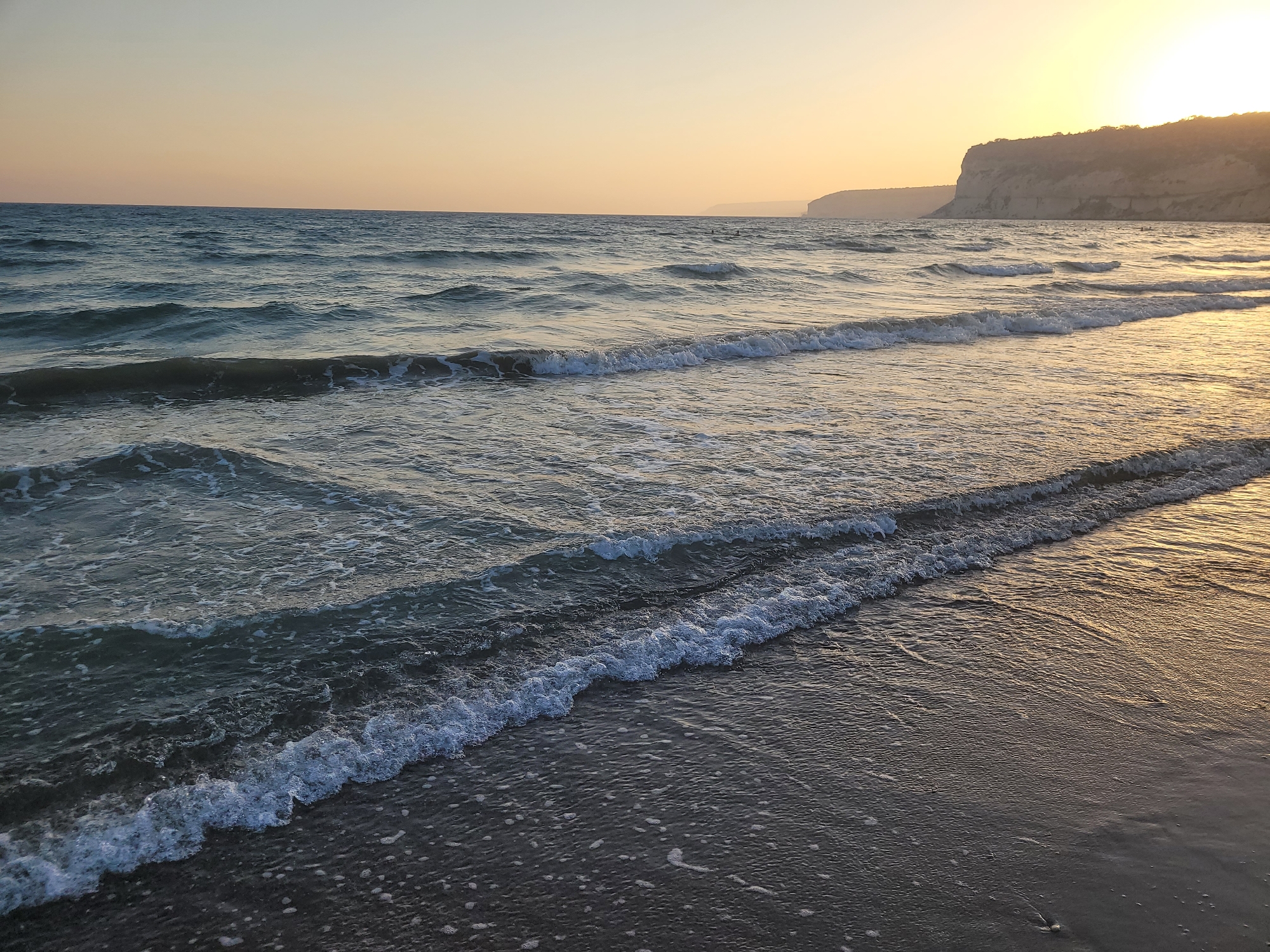 The image shows a serene seascape at Episkopi Beach, near Episkopi, Limassol, Cyprus, not in the United Kingdom. The sun is setting, casting a warm golden glow across the sky and reflecting on the calm sea. Gentle waves are rolling onto a sandy shore. In the distance, cliffs are visible along the coastline, silhouetted against the vibrant sunset. The scene is peaceful and tranquil, with no people or specific objects in sharp focus, emphasizing the natural beauty of the coastal landscape. The water is a deep blue-green, with white foam highlighting the crests of the waves. The sand is dark and wet, showing the texture left by the receding tide.