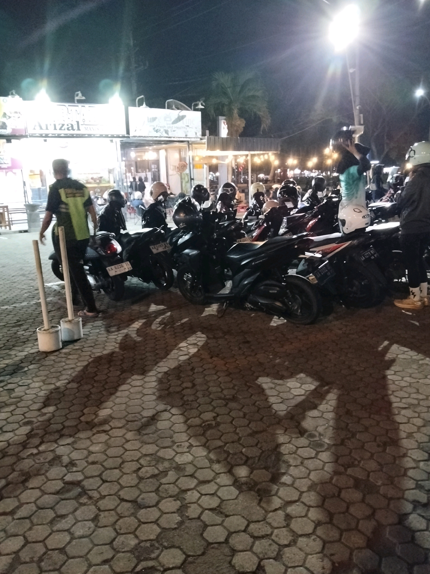 The image depicts a nighttime scene in front of a food stall area in Purworejo, Indonesia. In the foreground, two white cylindrical posts are visible on a hexagonal paver surface, casting distinct shadows. A person wearing a black shirt and a bright green safety vest is standing to the left of the posts, facing away from the camera. Behind this person and to the right, a collection of motorcycles is parked. Several individuals are seen near the motorcycles, some wearing helmets, suggesting they are either preparing to depart or have recently arrived. The background features illuminated signs for food stalls, with "Arizal MANIS" being prominently visible on one of the signs. The overall lighting suggests it is after dark, with artificial lights from the stalls and streetlights illuminating the area.