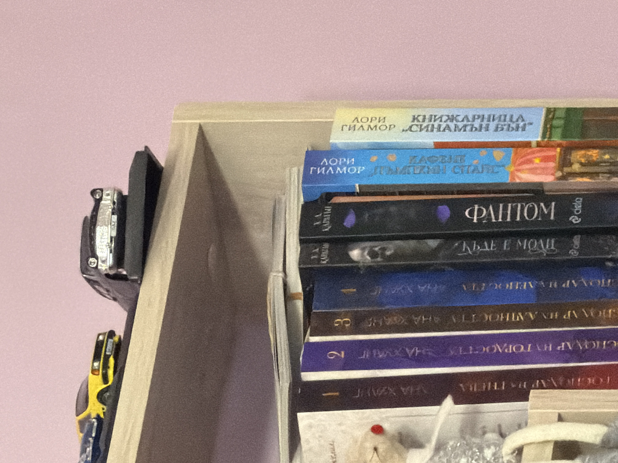 A close-up, eye-level shot showcases a wooden shelf unit against a light pink wall. The shelf is filled with books, their spines oriented towards the viewer. Two toy cars, one yellow and one black, are placed on top of the shelf. The yellow car is in the foreground, and the black car is positioned behind it. The books appear to be part of a collection, with some spines featuring numbers like "1" and "2". The spines of the books are predominantly dark in color, with some lighter accents. The lighting is even, and there are no visible shadows or glare. The background is a plain, textured pink wall. The image composition is focused on the shelf and its contents, with the top portion of the shelf and the toy cars being a significant element. There is no indication of the location being Plovdiv, Bulgaria.