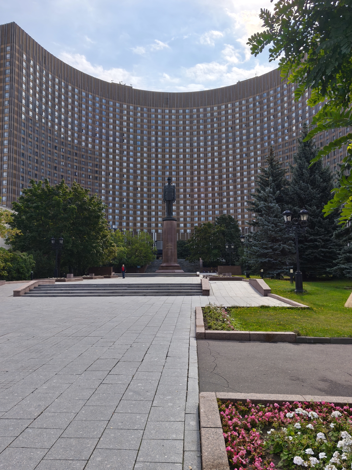 The image depicts the Cosmos Hotel in Moscow, Russia, a well-known landmark. In the foreground, a wide expanse of gray paving stones leads towards a set of wide steps. To the right of the steps, a meticulously manicured lawn borders a paved pathway. A dark lamppost stands on the lawn, and in the distance, a cluster of tall evergreen trees are visible. Further back, a large, curved building, the Cosmos Hotel, dominates the background. Its facade is adorned with numerous windows, arranged in a grid pattern. In front of the building, a statue of a man stands on a pedestal, with a single person in a red shirt walking near its base. The sky above is a mix of blue and white clouds. Several trees with green foliage are scattered throughout the scene, with some branches extending into the frame from the top right corner.