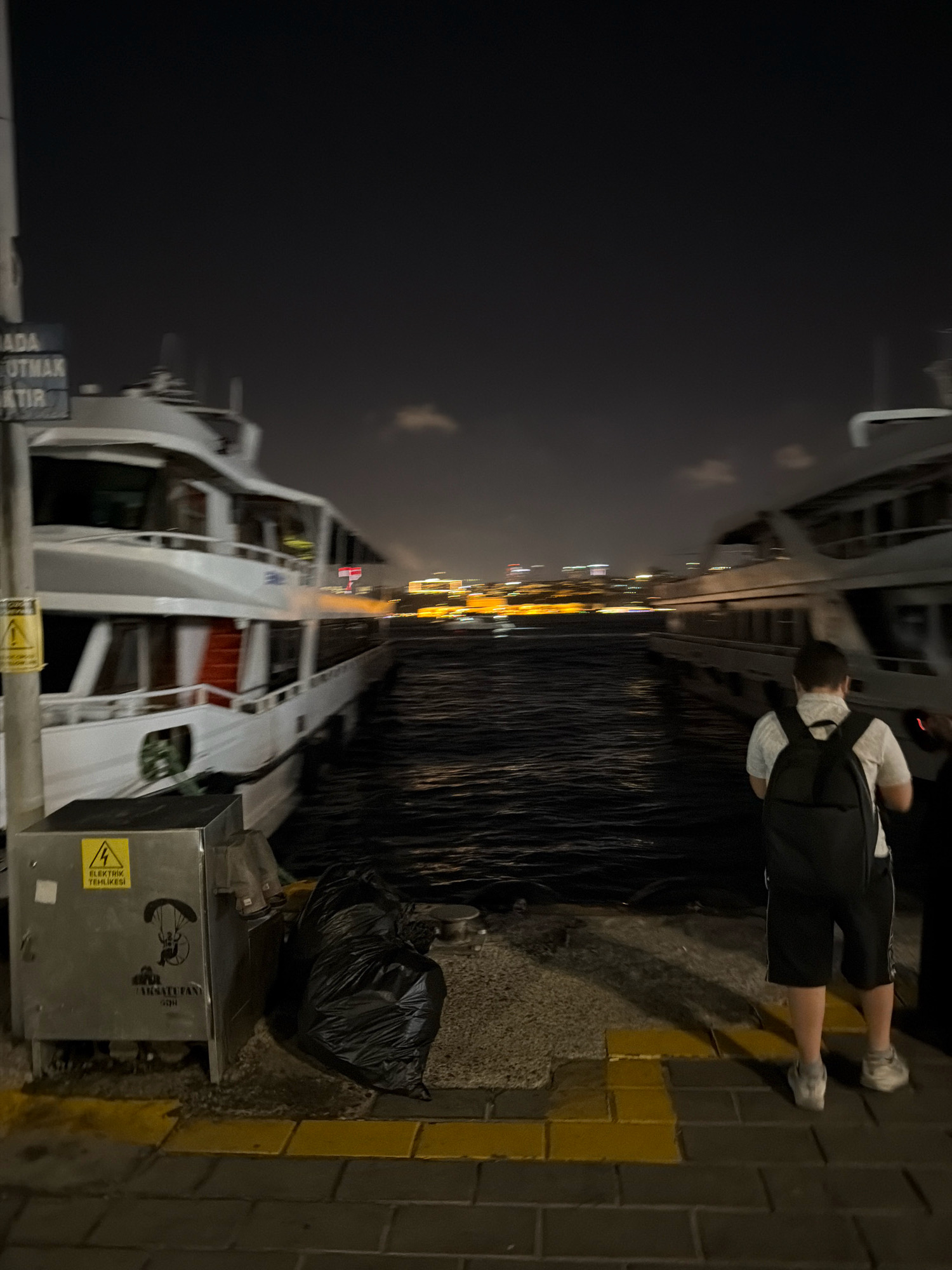 The image depicts a nighttime scene at a pier in Üsküdar, Turkey. A young person wearing a white shirt, black shorts, and a backpack is standing on the pier, facing away from the camera, looking towards the water. Two large ferries are docked on the left and right sides of the frame, their lights illuminating the dark water. The ferry on the left is white with red accents and has several windows. The ferry on the right is partially visible. The pier is made of brick pavers with yellow markings. To the left of the young person, there is a metal electrical box with warning signs and a sticker of a paraglider. A black garbage bag is leaning against the electrical box. In the background, the city lights of Istanbul are visible across the Bosphorus Strait. The overall atmosphere is that of a quiet, late-night port.
