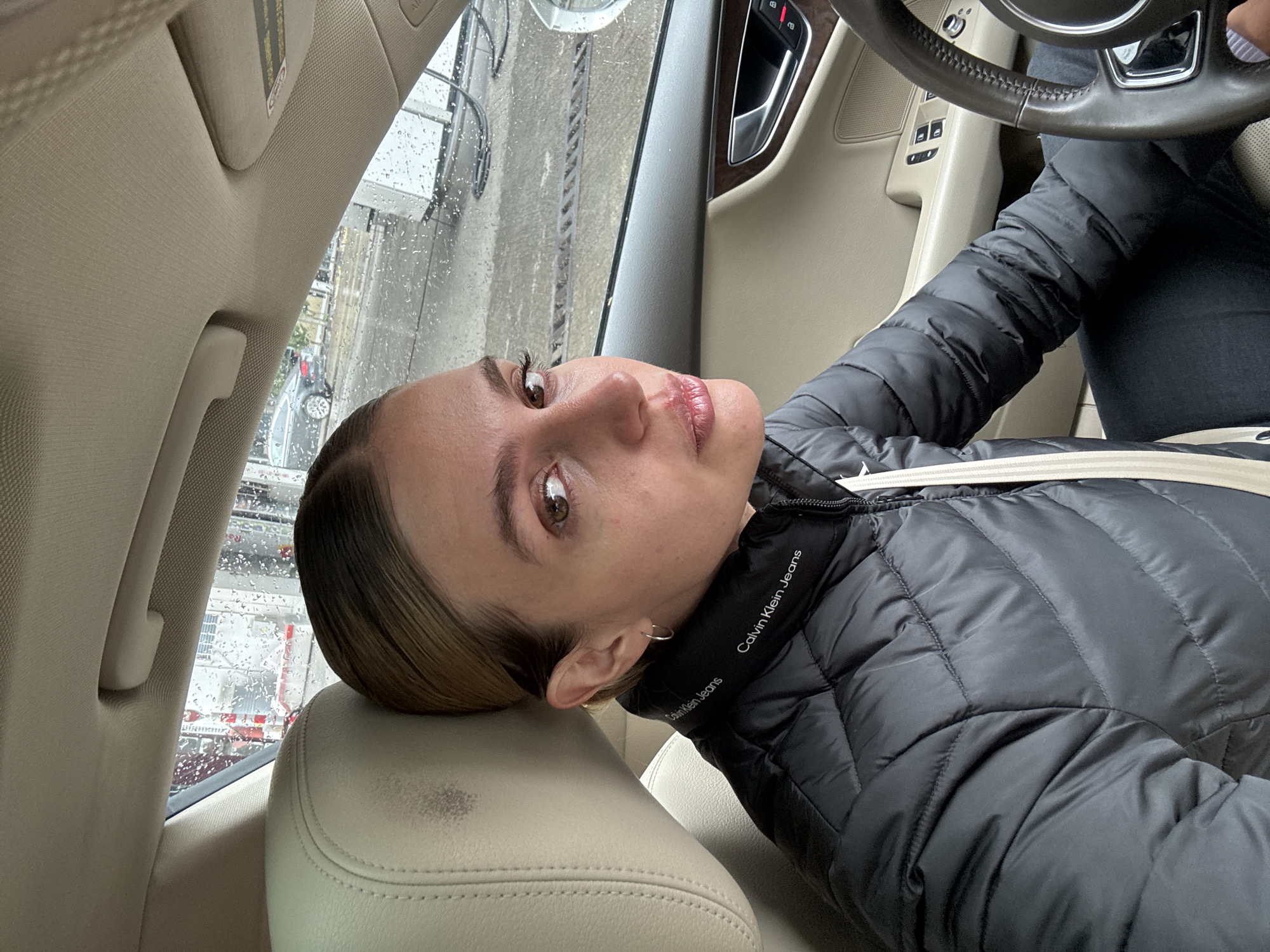 The image shows a woman taking a selfie from the driver's seat of a car in Tbilisi, Georgia. She is looking directly at the camera with a neutral expression. She has light brown hair, dark eyebrows, and brown eyes. She is wearing a black puffer jacket that features "Calvin Klein Jeans" text repeatedly on its collar. A light-colored seatbelt crosses her chest.
The car's interior is predominantly beige or tan, visible on the seats, roof lining, and door panels. Parts of the steering wheel, the driver's door with controls (including what appear to be window and mirror adjustments), and a door handle with a wood-like trim are discernible. A sun visor with an "AIRBAG" warning is visible on the ceiling. There is some minor discoloration or wear on the driver's seat headrest.
Outside the car, the window is covered in water droplets, indicating either rain or that the vehicle is in a car wash. Through the wet window, blurred industrial structures or components of an automatic car wash are visible, along with a partial view of another car. The activity appears to be a moment of pause, possibly during a car wash or while parked in rainy conditions. The lighting suggests it is daytime.