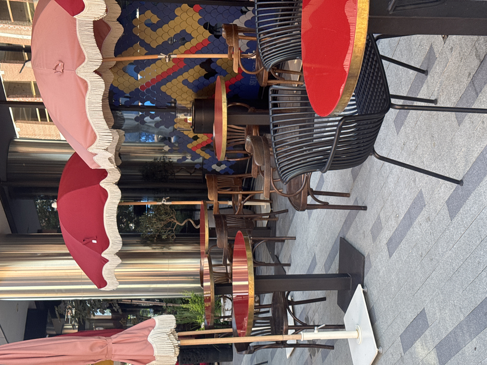The image depicts an outdoor seating area of a restaurant or cafe. There are several tables with red tops and gold trim, accompanied by various styles of chairs. Some chairs are dark wood with a classic design, while others are modern black metal with a slatted back. Two large, decorative umbrellas, one pink and one red, provide shade. In the background, a building with large windows and a colorful, patterned facade is visible. The ground is paved with grey stone tiles. The scene is devoid of any people. The image does not provide any specific clues about its location in Yekaterinburg, Russia.