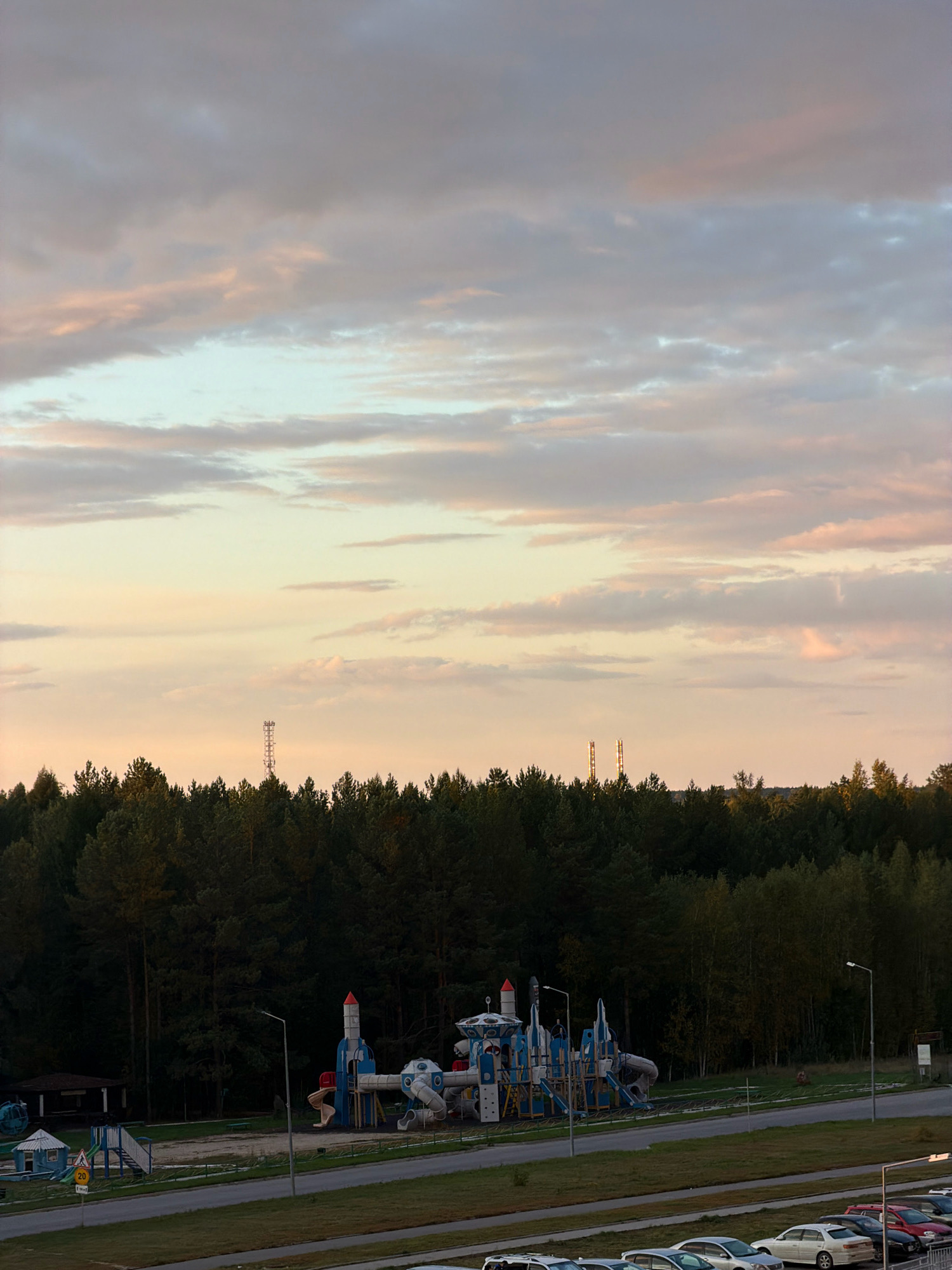 The image depicts an outdoor scene at dusk in the city of Tsiolkovsky, Russia. In the foreground, a row of parked cars is visible on a paved surface. A street with a grass median separates the parking area from a playground. The playground features several abstract, brightly colored structures, including what appear to be rocket-shaped elements and tubes, suggesting a space or futuristic theme. Beyond the playground, a dense forest of dark green trees forms a backdrop. In the sky, a mix of soft, muted clouds with hints of pink and yellow streaks suggests the setting sun. Two tall cell towers are visible above the tree line in the distance. The overall atmosphere is calm and serene.