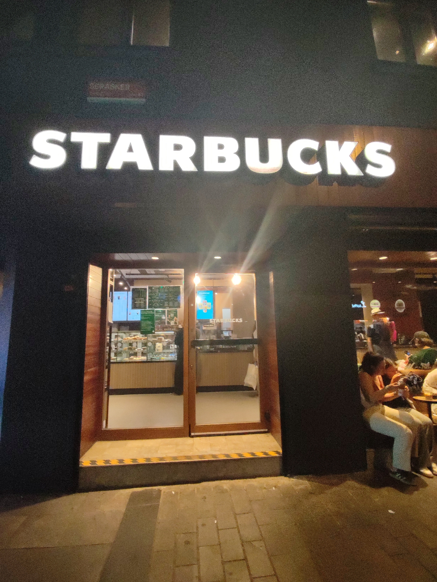 The image shows the exterior of a Starbucks coffee shop at night. A large, illuminated sign with "STARBUCKS" in white lettering is visible above the entrance. The entrance consists of two glass doors framed by dark wood. Inside, the coffee shop appears well-lit, with displays of food items and menus visible on the walls. Several people are seated outside on a bench, some of whom are engaged in conversation or looking at their phones. The sidewalk in front of the shop is paved with bricks. A small sign with red lettering is visible above the Starbucks sign. The overall impression is of a busy urban environment at night. The location is identified as Kadıköy, Turkey.