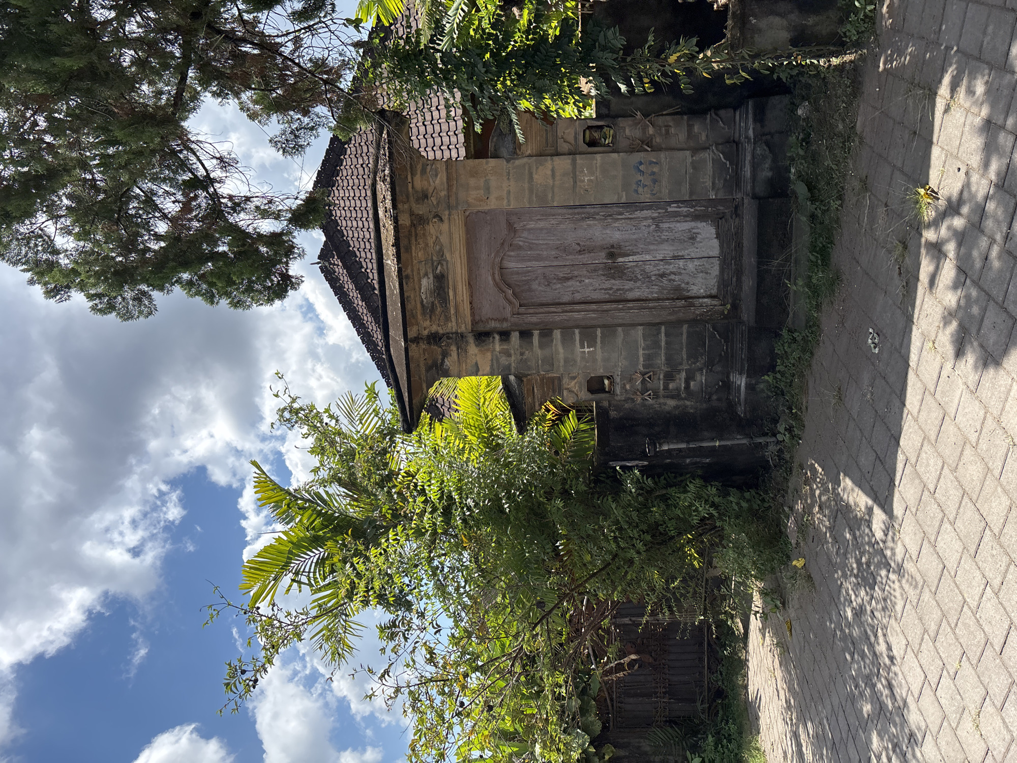 The image captures a weathered, traditional stone or concrete structure, possibly a shrine or an old building entrance, standing under a bright blue sky dotted with white cumulus clouds. The building features a dark, reddish-brown tiled roof and a prominent, old wooden door or gate with a distinctive carved top and visible signs of age. The stone facade below the roof shows faded decorative patterns and small recessed openings.
Lush, green tropical vegetation, including tall coniferous-like trees, broad-leafed plants, and palm-like fronds, densely surrounds and partially obscures the structure, suggesting a rich natural environment. Sunlight illuminates the scene, casting distinct shadows from the trees and the building onto a paved pathway made of interlocking grey blocks that runs along the right side of the structure. Some grass and weeds sprout from the cracks in the paving and at the base of the wall.
The scene depicts a tranquil, static moment on a sunny day, likely morning or early afternoon, given the bright light and shadow patterns. No people or specific activities are visible. The architectural style and abundant tropical foliage are consistent with the location context of Kuta Selatan, Indonesia. No discernible text is visible in the image.