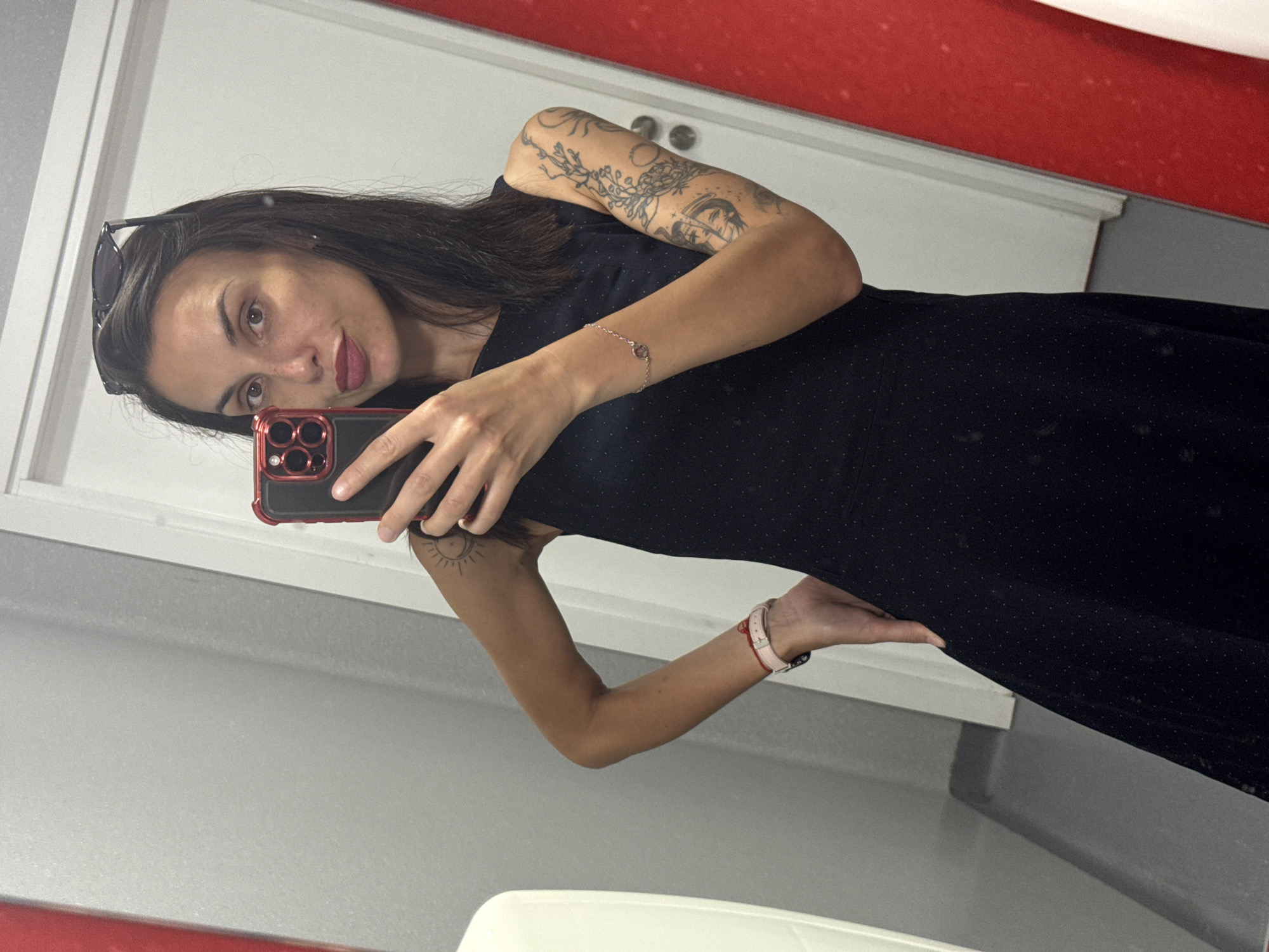 A woman with dark hair and a fair complexion is taking a selfie in a bathroom mirror. She is wearing a black, dotted dress and sunglasses on her head. Her left arm, fully visible, has multiple tattoos, including floral designs and other intricate patterns, extending from her shoulder to her wrist. A delicate bracelet adorns her left wrist. Her right arm, also visible, has a small sun tattoo and is wearing a light pink watch. She is holding a red smartphone with a black screen. The background reveals a modern bathroom interior with white and gray surfaces, a red countertop along the top of the mirror, and part of a white toilet seat visible at the bottom. Without more information, the exact location in Pleven, Bulgaria cannot be determined.