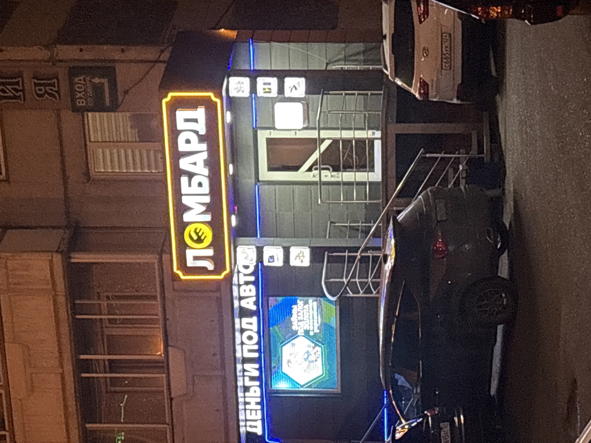 A nighttime shot shows a building facade in Krasnoyarsk, Russia. The sign "ЛОМБАРД" (Lombard, or Pawnshop) in neon yellow is prominently displayed. The sign below reads "ДЕНЬГИ ПОД АВТО" (Money for Cars). To the side and above the door are the symbols for money, watches, and jewels. Two cars are parked along the curb to the right. The license plate on the white car reads "С685УК 124." A black sedan is visible in front of the white car. Another sign reads "ВХОД СО ДВОРА" (Entrance from the Courtyard) and has a left-pointing arrow.