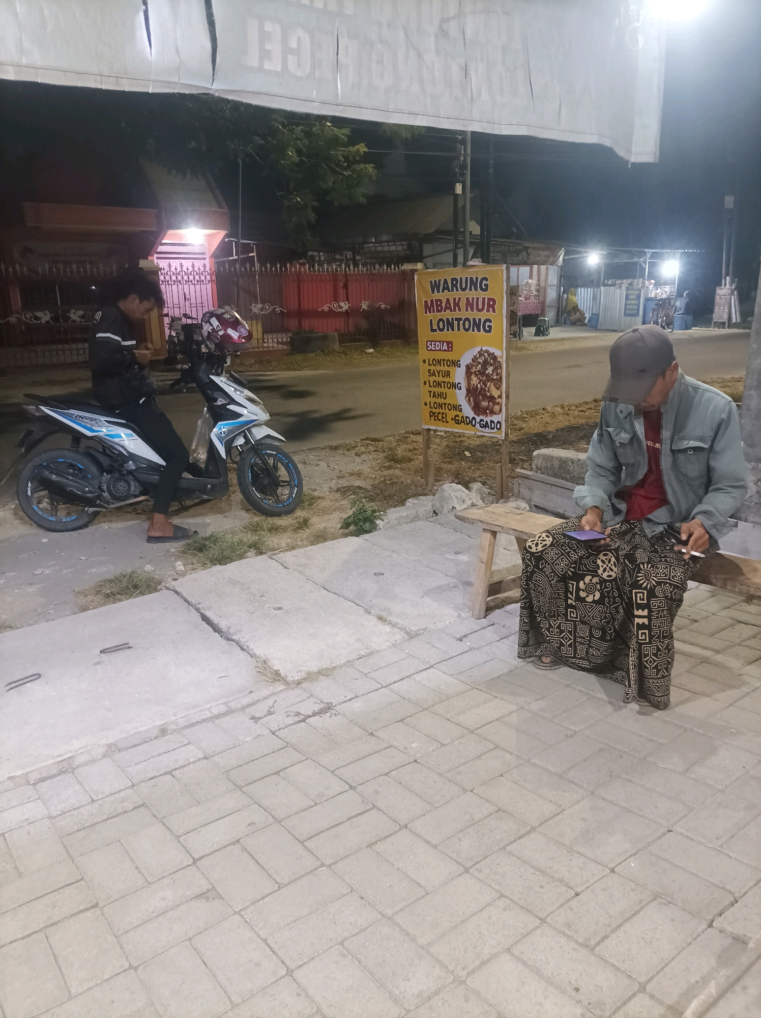 The image depicts a street scene at night in Sumuragung, Indonesia, near a food stall. In the foreground, a man wearing a grey jacket, a red t-shirt, a grey baseball cap, and a sarong with a black and beige patterned print is seated on a wooden bench. He is holding a smartphone in his right hand and a cigarette in his left hand. To his left, a young man wearing a dark jacket with white stripes and black pants is sitting on a white and blue motorcycle. He is looking down at his phone. A yellow sign for "WARUNG MBAK NUR LONTONG" is visible behind the motorcycle. The environment consists of a paved sidewalk, a street, and a building with an illuminated sign in the background. The overall lighting suggests it is nighttime.