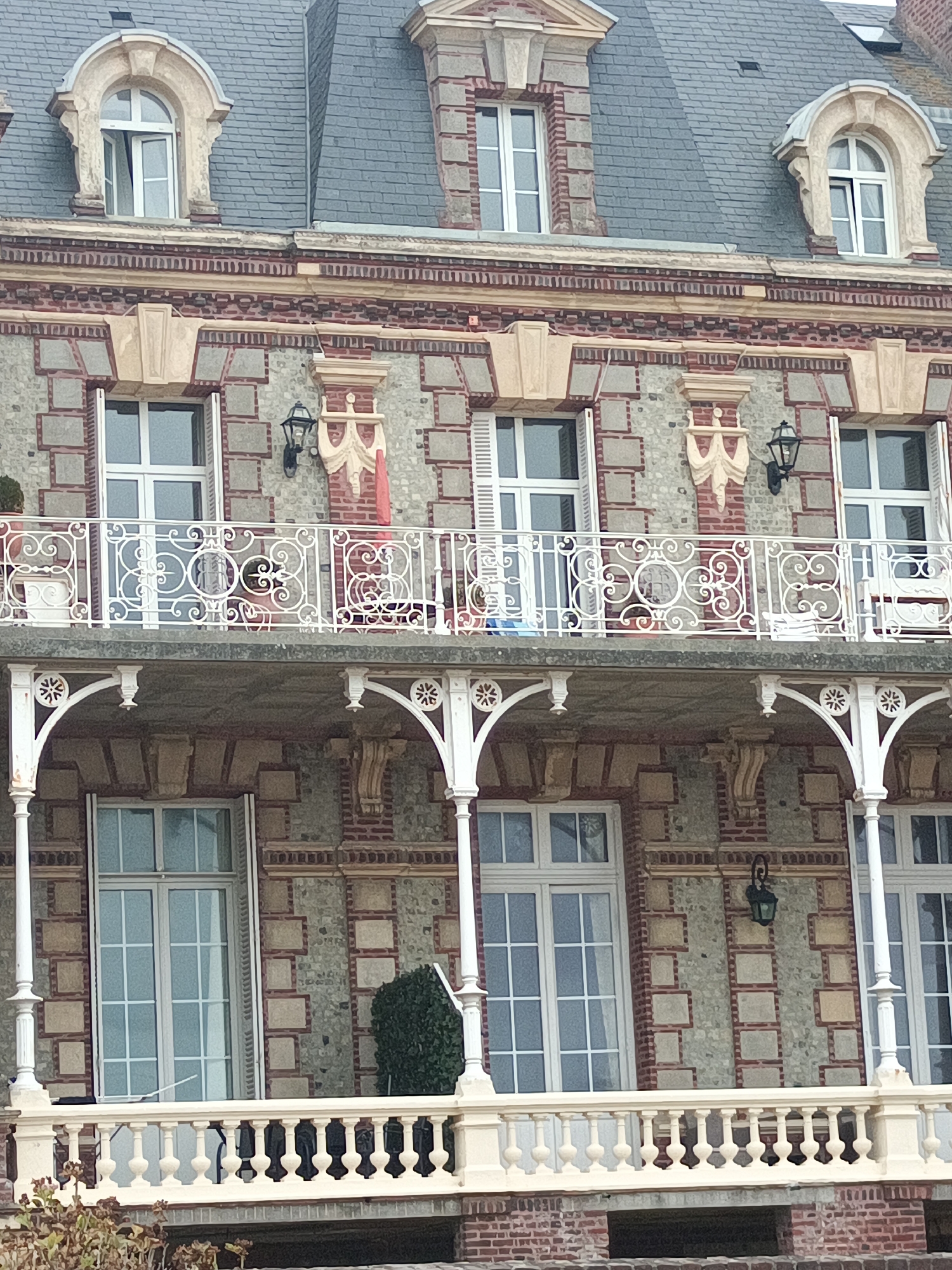 A facade of a multi-story building in Houlgate, France, is shown. The architecture is characterized by a patterned brick and stone exterior, featuring numerous windows with white frames and shutters. Several white balconies with ornate railings and decorative supports are visible. One balcony on the upper level has a white metal railing with intricate scrollwork. The building has a grey tiled roof with dormer windows. The lower part of the image shows more brickwork and a glimpse of dry vegetation in the foreground.
