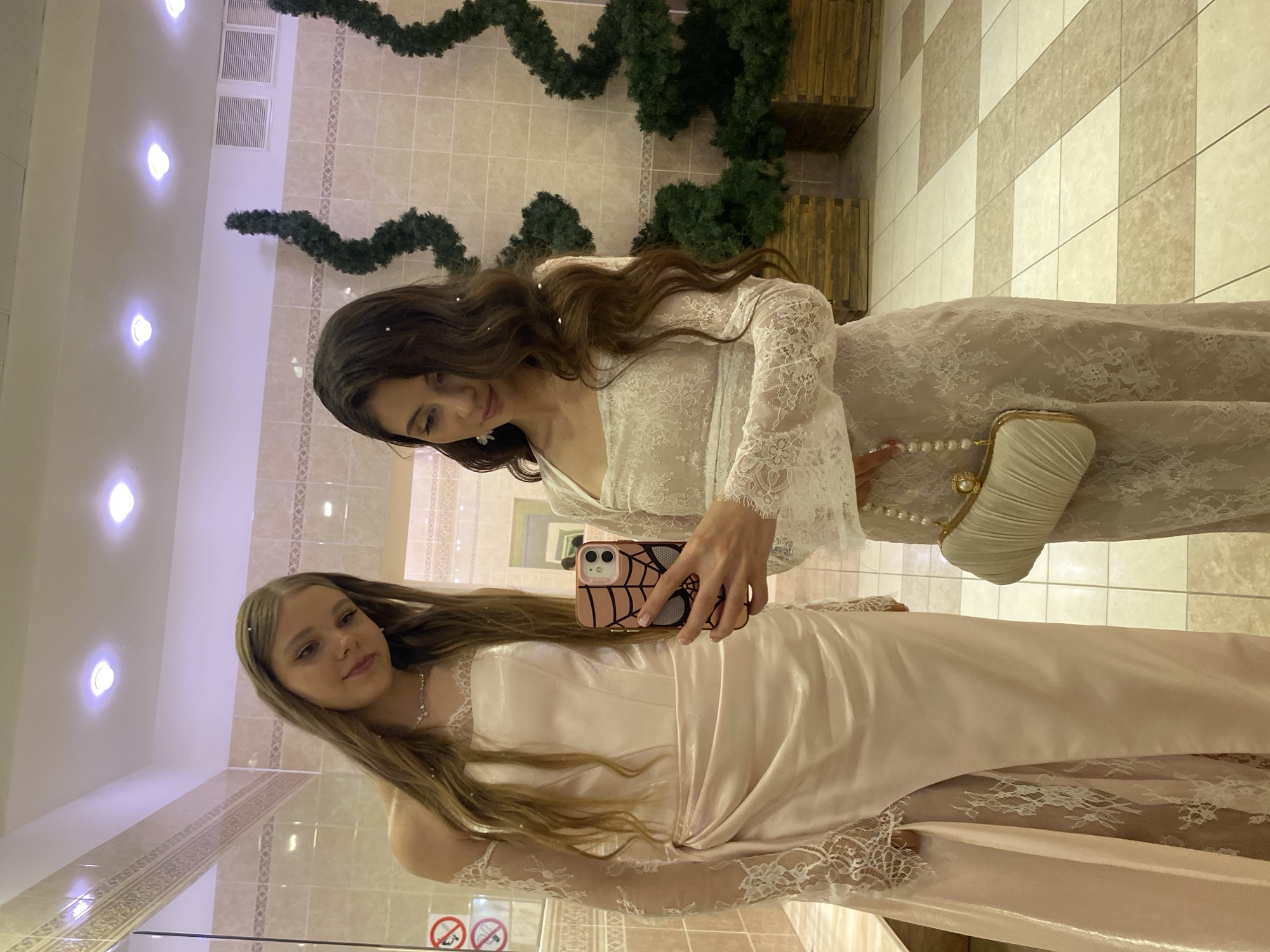 This media file captures a mirror selfie of two young women in formal attire. The woman in the foreground, on the left, is wearing a light pink satin dress with lace detailing on the sleeves and hem. She has long, light brown hair and is looking towards the camera with a gentle smile. The woman in the background, on the right, is wearing a white lace dress. She has long, dark brown hair and is also looking at the camera. She is holding a smartphone in her right hand, which is angled to capture the reflection.
Both women appear to be dressed for a special occasion, possibly a formal event or celebration. The background suggests an indoor setting, likely a room with tiled walls and floors. Decorative evergreen garlands are visible in the upper part of the image, suggesting a festive atmosphere, possibly around a holiday season like Christmas or New Year. The lighting appears to be artificial, with bright circular lights in the ceiling.
The overall mood of the image is cheerful and celebratory. The women seem happy and are posing for a photograph. The presence of mirrors indicates they might be getting ready in a dressing room or a similar space. There are no specific indicators of time of day or weather conditions, as it is an indoor shot. There is no visible text in the image, except for some pictograms on signs in the background, which are too small to be legible. The location context provided is Votkinsk, Russia.