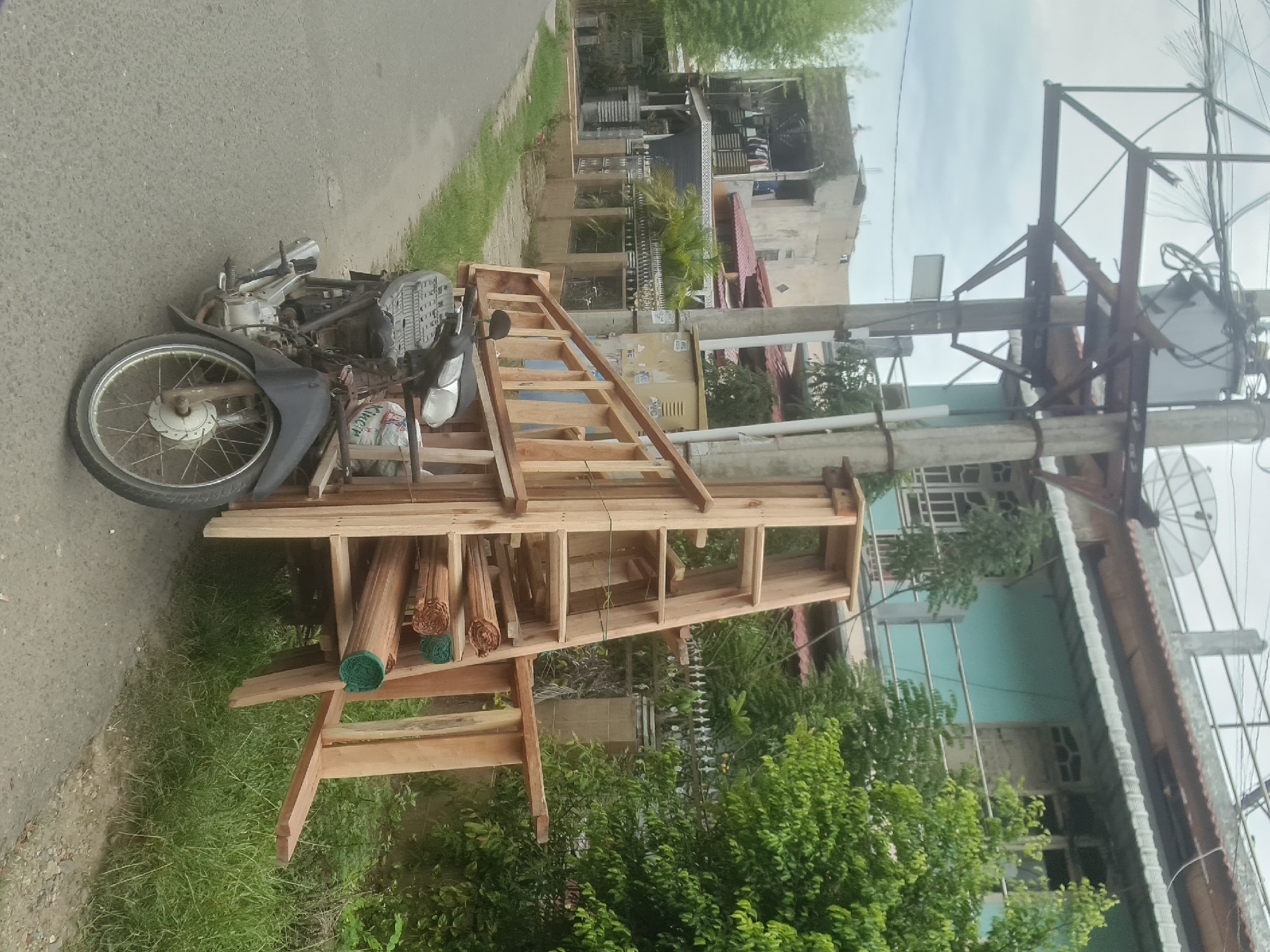 A wooden ladder and other lumber are precariously loaded onto the back of a black motorcycle, parked on the side of a paved road in Lhokseumawe, Indonesia. The motorcycle is positioned on the left side of the frame, with its front wheel facing the viewer. The ladder, constructed from light-colored wood, is significantly taller than the motorcycle and extends out to the right. Several rolls of what appear to be woven mats or reeds are stacked horizontally within the ladder structure. The environment suggests a residential or semi-urban area, with buildings and greenery visible in the background. A utility pole with electrical wires is prominent in the upper portion of the image. The sky is overcast.