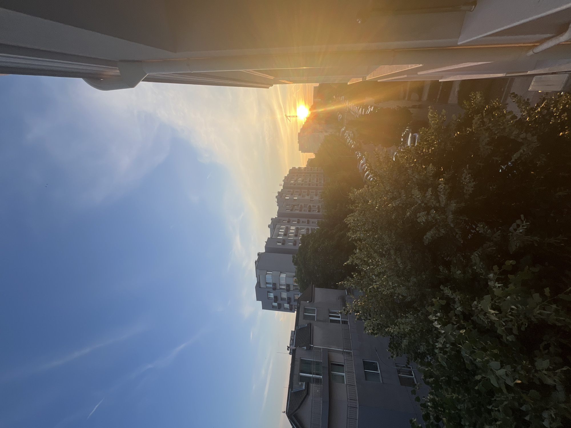 The image captures a sunset over a residential area in Burgas, Bulgaria. In the foreground, a section of a building with a white downspout is visible on the right. Beyond this, lush green trees dominate the middle ground, partially obscuring the view of apartment buildings. These buildings are several stories high and have a modern architectural style with numerous windows. A street lined with parked cars runs between the buildings. The sky is a clear blue with thin, wispy clouds, and the setting sun casts a warm, orange glow across the scene, creating lens flare effects.