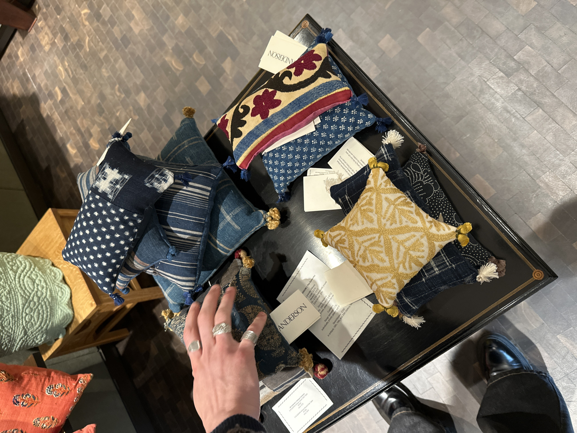 This overhead shot captures a retail display featuring an assortment of decorative cushions and textiles on a dark, possibly lacquered, table with a gold trim. A person's right hand, adorned with several silver-toned rings, reaches towards a cushion, indicating engagement with the items. The person's feet, wearing black shoes and dark jeans, are visible in the bottom right corner, standing on a light grey tiled floor.
The cushions vary in size and pattern, showcasing a range of designs including intricate floral embroidery in gold and red on beige, geometric patterns in shades of blue and white, and striped fabrics. Many of the cushions are accented with small tassels at the corners. White price tags and information cards are interspersed among the items.
Visible text on the cards includes the brand name "ANDERSON" and "ERSON x The Dyeworks." Products listed include "Lavender Pillows" with prices like "Medium: £315," "Large: £375," and "Extra Small: £90." "Dinner Napkins" are priced at "£35/£150 individually priced" and "£95-£800 Set of 6/8/12." "Cushions" are described as "Range between £275-" and "Individually priced," and "Sketchbooks £200" are mentioned as being "in antique, 18th, 19th, and 20th Century." This detail confirms the setting as a high-end retail environment or a gallery in London, United Kingdom.
A portion of a wooden table and a light green, circular quilted mat are partially visible on the left side of the frame, suggesting other display elements nearby. The overall ambiance is refined, typical of a boutique showcasing artisan or luxury home goods.