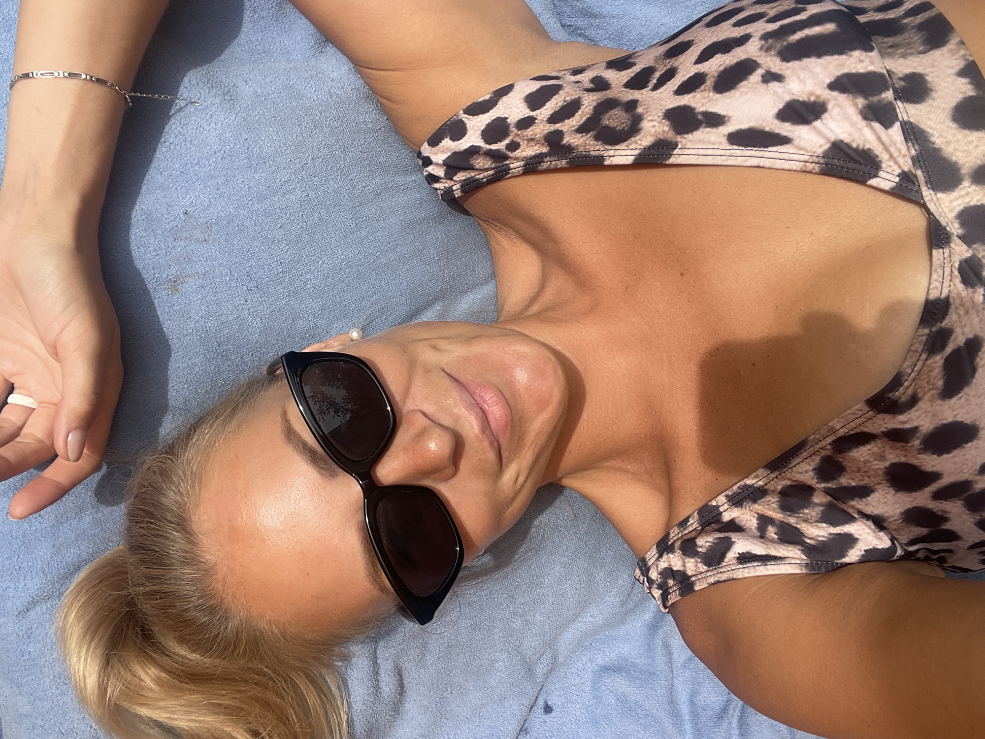 A fair-skinned woman with blonde hair pulled into a ponytail is lying on a blue towel, viewed from a high-angle perspective. She is wearing black cat-eye sunglasses and a leopard-print bikini top. Her left arm is outstretched above her head, and her right arm is bent with her hand near her head. A silver bracelet is visible on her left wrist. The environment is outdoors, as suggested by the lighting and shadows. The image does not contain any identifiable features of the city of Baranovka, Russia.