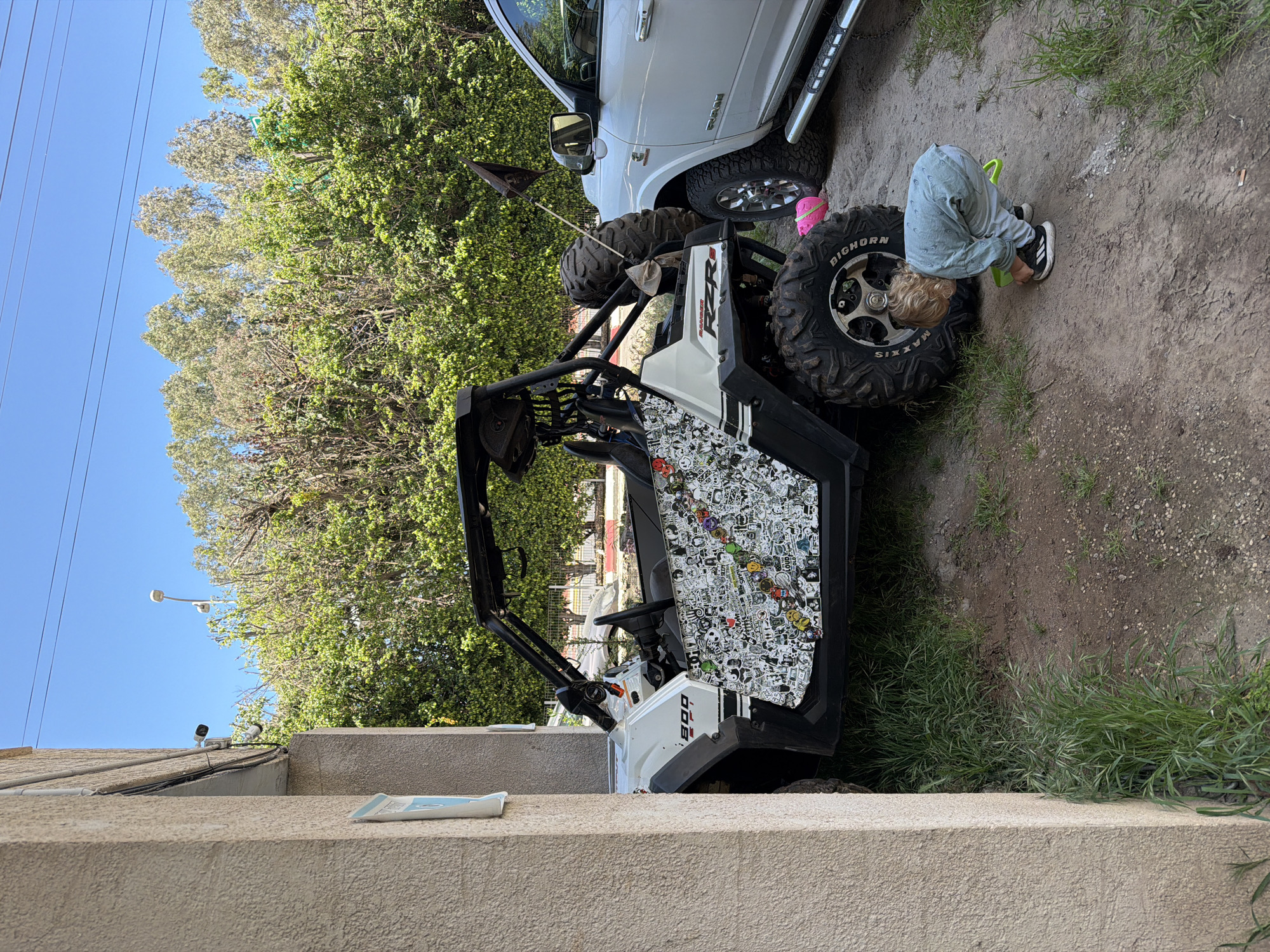This image, taken in Kiryat Motzkin, Israel, depicts an outdoor scene on a bright, clear day. The primary subject is a white Polaris RZR 800 utility terrain vehicle, parked on a dirt and gravel surface. The RZR features "RZR 800" branding and its side panel is extensively covered with a sticker bomb pattern of black and white graphics, interspersed with small colorful accents. It has large, rugged "MAXXIS BIGHORN" tires, and a small black flag on a pole is attached to its rear.
Next to the RZR, a young child with blond hair, dressed in a light-colored long-sleeved shirt, jeans, and black and white sneakers, is bending over. The child appears to be playing or examining something near the vehicle, holding a small green object. A white Dodge Ram pickup truck is parked behind the RZR, with its "RAM" logo visible on the side and a pink bucket near its wheel.
The background features a line of green trees against a clear blue sky, with power lines visible in the upper left. In the immediate foreground, a light-colored, textured building wall partially frames the scene. Surveillance cameras are visible on a rooftop in the distance. The ground is a mix of dirt and sparse grass, suggesting an unpaved outdoor area. The overall impression is of a casual, daytime scene outdoors.