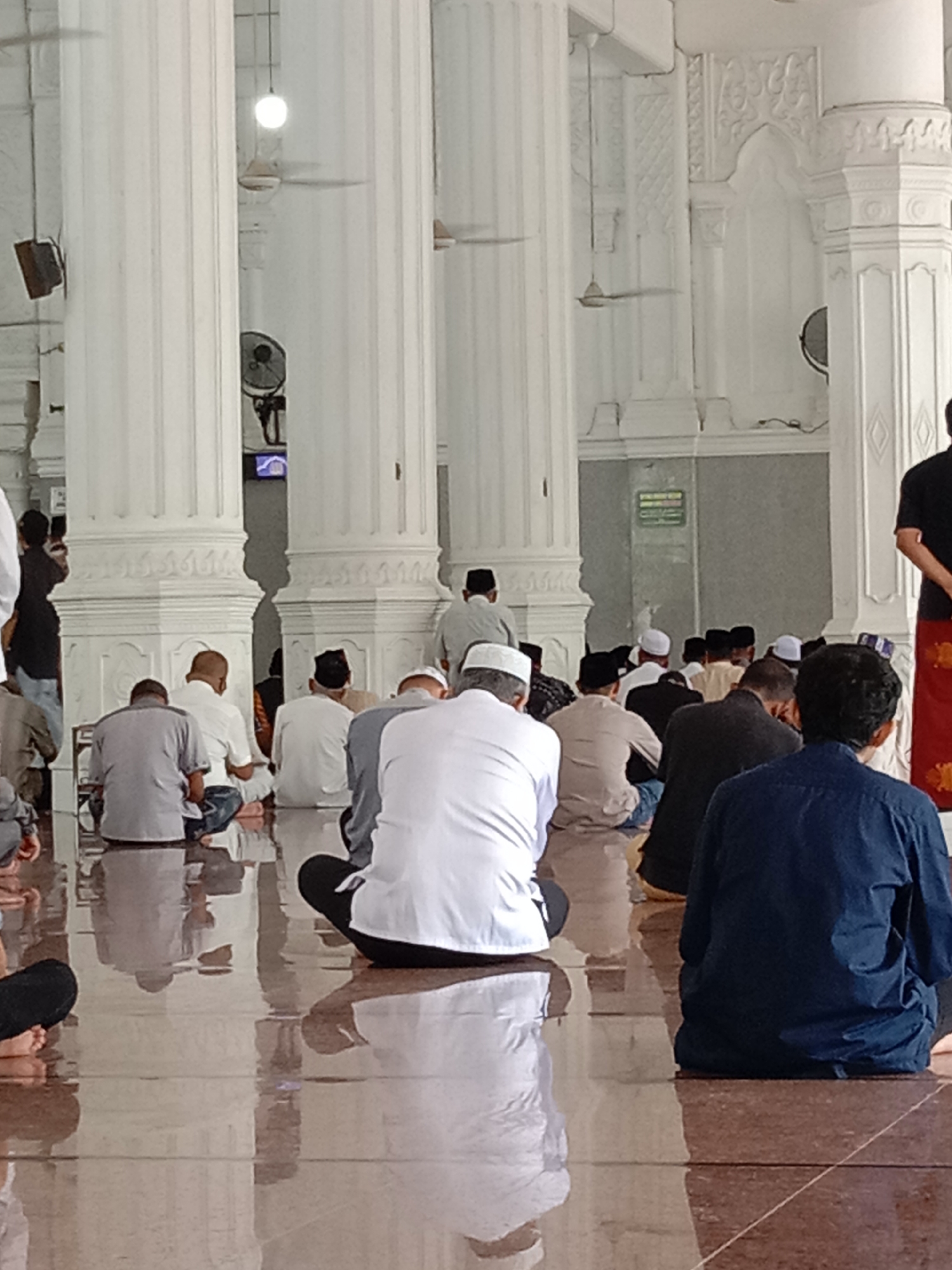 The image shows the interior of a mosque with many men sitting on the floor in prayer. The mosque appears to be large and well-maintained, with tall white columns and a polished floor that reflects the light. The men are dressed in traditional Islamic attire, and their faces are not visible as they are either looking down or their backs are to the camera. The lighting suggests it is daytime, possibly with natural light coming from windows or openings not visible in the frame. The atmosphere seems to be one of solemnity and devotion. The location is identified as the Baiturrahman Grand Mosque in Lhokseumawe, Indonesia, a significant religious landmark.