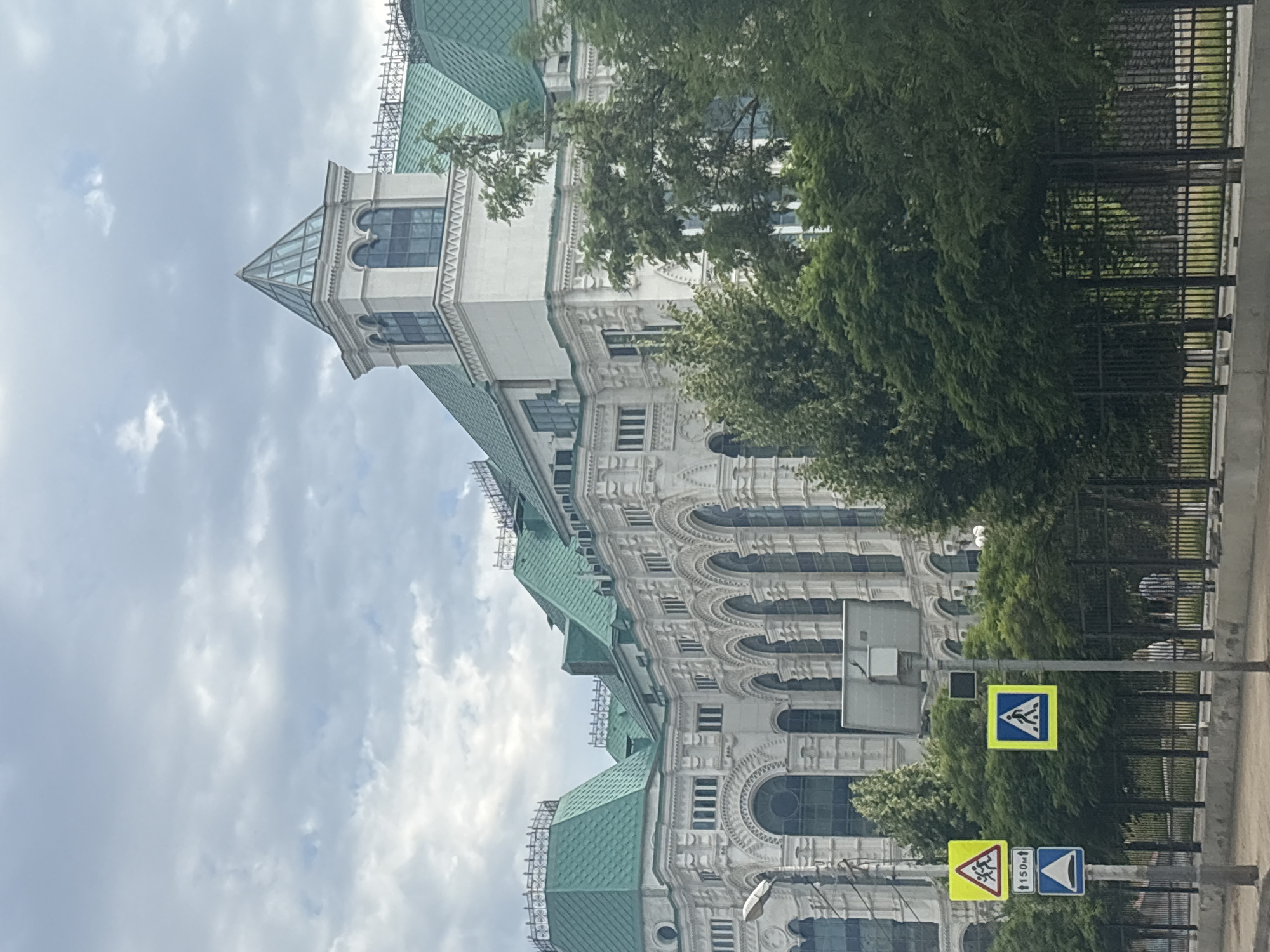 The image depicts a grand, ornate white building with a distinctive green roof, likely a government or cultural institution, located in Astrakhan, Russia. The architecture features multiple arched windows and intricate decorative elements. In the foreground, a street scene is visible with traffic signs, including a pedestrian crossing sign and a children crossing warning sign. Lush green trees line the front of the building and a black metal fence separates the property from the street. The sky is overcast with clouds. There are no people clearly visible in the image, though the context of a city street suggests their presence.