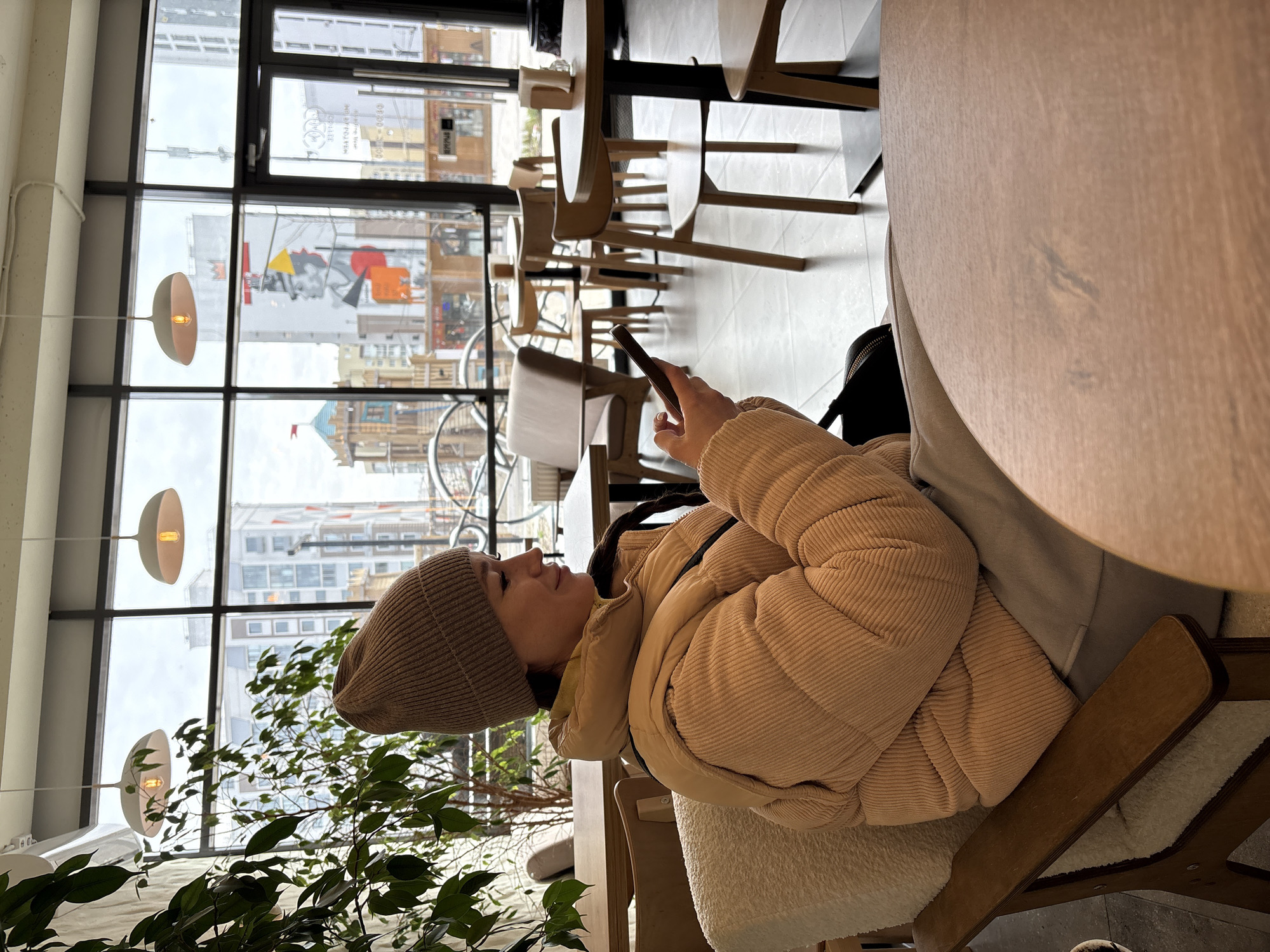 A woman is seated in a chair indoors, looking at her mobile phone. She is wearing a tan beanie and a tan corduroy puffer jacket. A black backpack is near her. There are tables and chairs visible in the background. Through a window, buildings can be seen. Based on visible signage and architectural styles, the location is likely in Ulyanovsk, Russia. The setting appears to be a cafe or restaurant. A green potted plant is in the foreground.