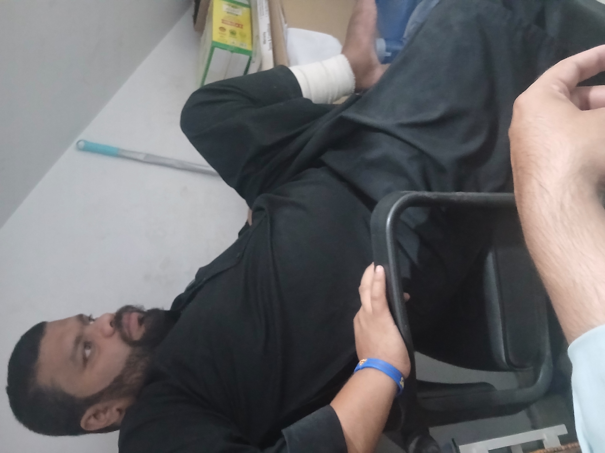 The image shows a medium shot of a man sitting in a black office chair. He is wearing a black shalwar kameez. He has a beard and is looking to his left. His right arm is resting on the armrest of the chair, and he is wearing a blue wristband. His left leg is bent at the knee, and his foot is bandaged. In the background, a mop with a blue handle leans against a white wall. To the right of the mop, there are some stacked boxes, one of which is yellow and green. The man appears to be in a room with a sloped ceiling. The scene is not clearly identifiable as being in Peshawar City Tehsil, Pakistan, though the cultural attire of the man suggests a South Asian context.