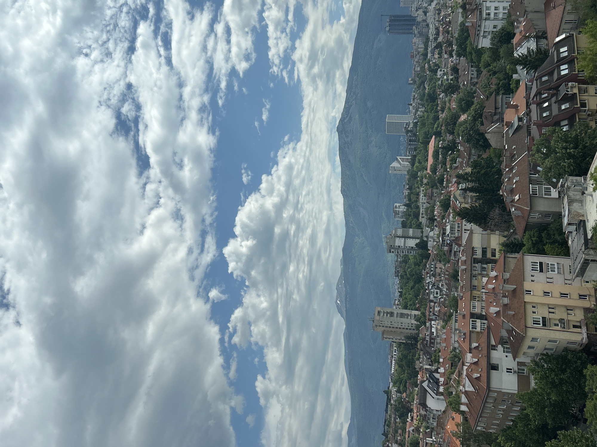 The image displays a panoramic daytime view of a bustling city nestled against a majestic mountain range, identifiable as Sofia, Bulgaria, with Vitosha Mountain in the background.
The upper portion of the frame is dominated by a bright blue sky, scattered with numerous white, fluffy clouds that vary in size and shape, indicating clear and fair weather. Below the sky, the imposing mountain stretches across the midground. Its slopes appear dark green, covered in dense vegetation, with hints of higher, possibly rocky or less vegetated areas towards the peaks.
In front of the mountain, the urban landscape of Sofia unfolds, occupying the foreground and midground. It features a dense collection of buildings, primarily residential, with many structures showcasing classic European architecture, characterized by red-tiled roofs and multiple stories. Green trees are interspersed throughout the city, providing a natural element amidst the urban sprawl. A few taller, more modern high-rise buildings are also visible, contrasting with the traditional architecture.
The scene captures the city under bright daylight, likely late morning or early afternoon, with strong illumination highlighting the architectural details and the natural environment. There is no specific human activity or event depicted, nor is any text visible within the frame. The image beautifully illustrates the close relationship between the vibrant city of Sofia and the natural beauty of Vitosha Mountain.