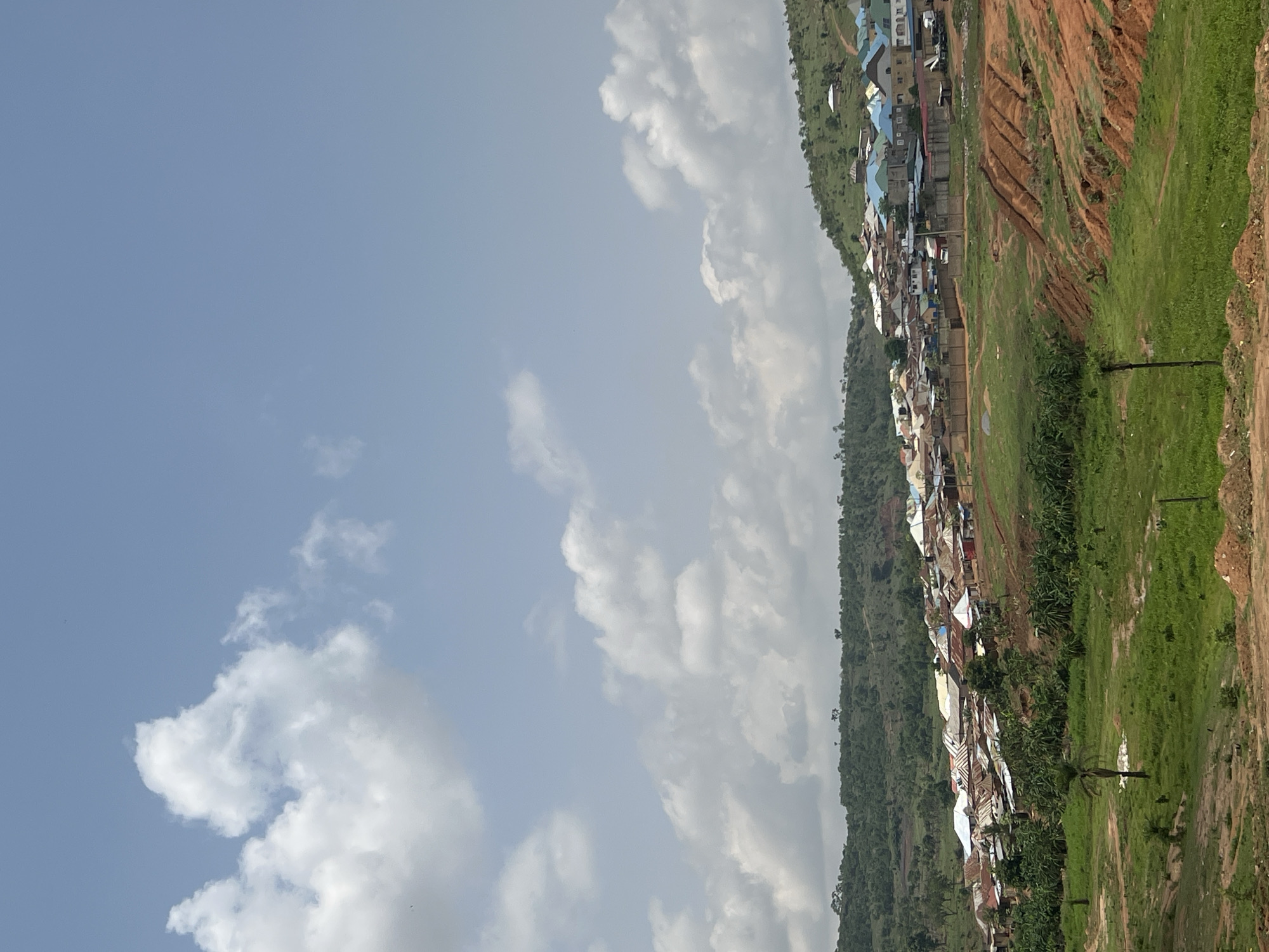 The image depicts a hillside settlement, characterized by tightly packed dwellings with varied roofing materials including metal sheets, situated along a green, sloping terrain. Visible ground erosion marks the area. The background is dominated by a partly cloudy sky. The scene is likely located in a hilly or peri-urban area in Abuja, Nigeria.