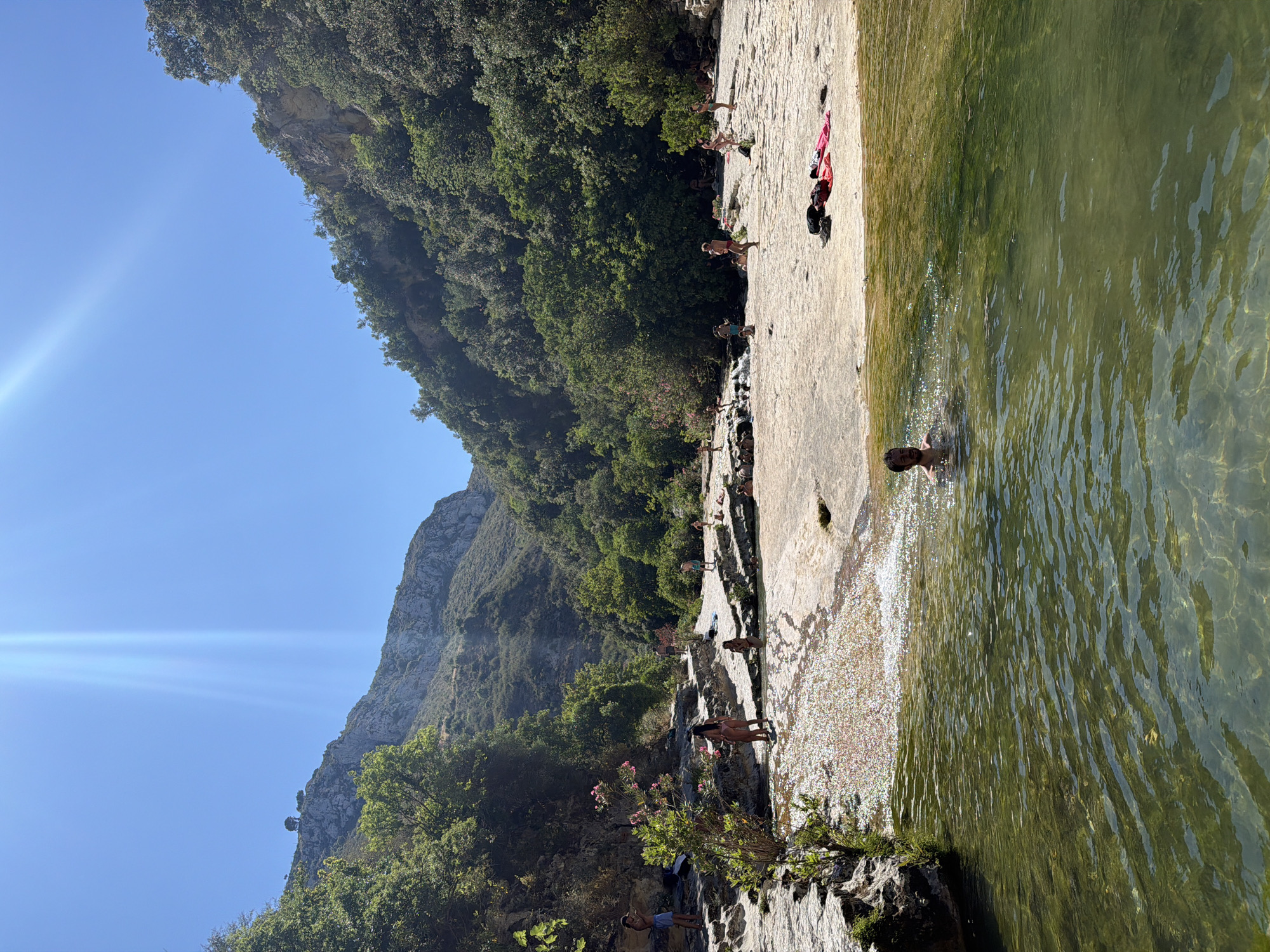 The image depicts a natural swimming area with a shallow river flowing over a rocky bed. Several people are visible, some swimming in the river and others relaxing on the sun-drenched rocky banks. The surrounding environment is lush and green, with trees and foliage covering the steep hillsides that form a gorge. The sky is clear and blue, with the sun casting bright rays, creating a vibrant and inviting atmosphere. Based on the provided context that this location is in the city of Noto, Italy, this scene is likely the Calamosche beach or a similar natural pool within the Vendicari Nature Reserve, known for its pristine beauty and crystal-clear waters.