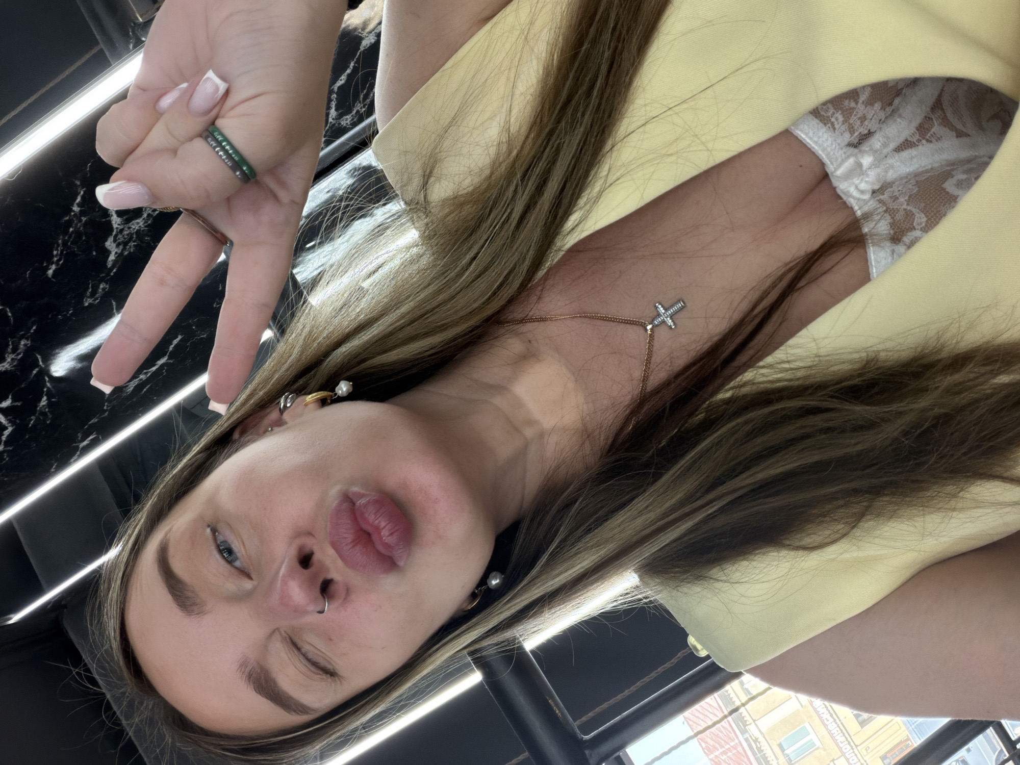 A woman is posing for a selfie. She is wearing a yellow blazer and a white lace top underneath. She has a nose ring and multiple earrings. She is winking and making a peace sign with her left hand. A gold chain with a cross pendant hangs around her neck. The background includes a black marble-like surface and what appears to be an urban environment with buildings, possibly in Moscow, Russia.