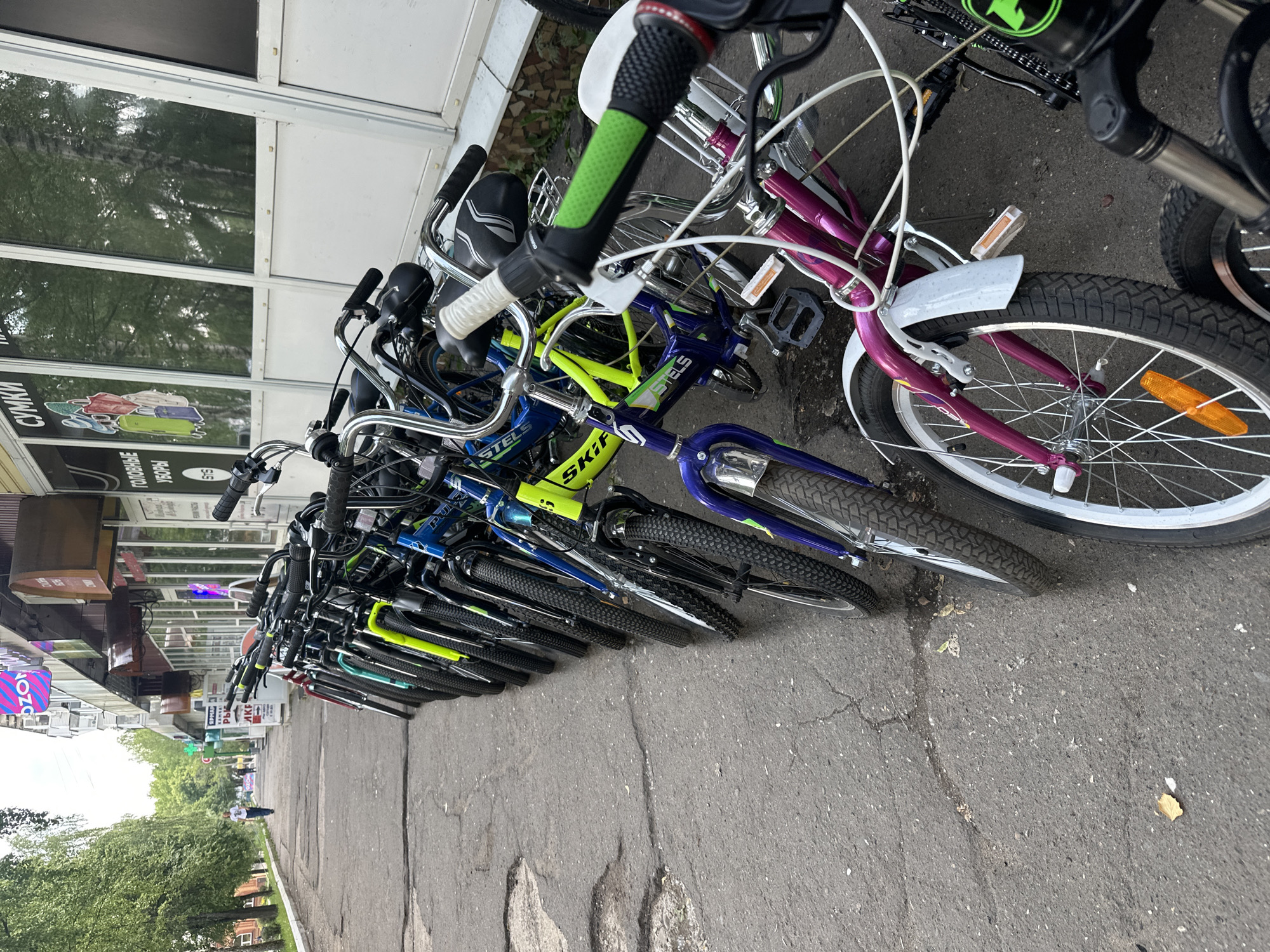 A line of bicycles is parked along a cracked asphalt sidewalk in front of a commercial building. The bicycles are of various colors, including blue, yellow, and purple, and some have visible brand names like "STELS." The building behind the bicycles has large windows and signs with Russian text, indicating it is a shop. Trees line the sidewalk to the left of the scene, and a person is walking in the distance. The weather appears to be overcast.
