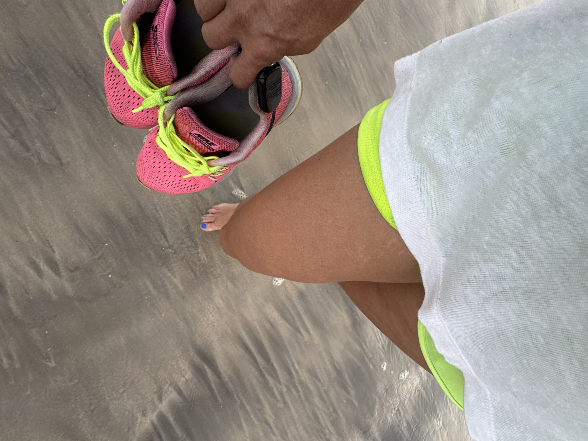 A person stands at the edge of the beach in Jimbaran, Indonesia, holding a pair of pink and neon-yellow running shoes. The person wears a white t-shirt and neon-yellow shorts. Their right foot is visible, with blue nail polish. The sand is wet and reflective, indicating the shoreline of the ocean.