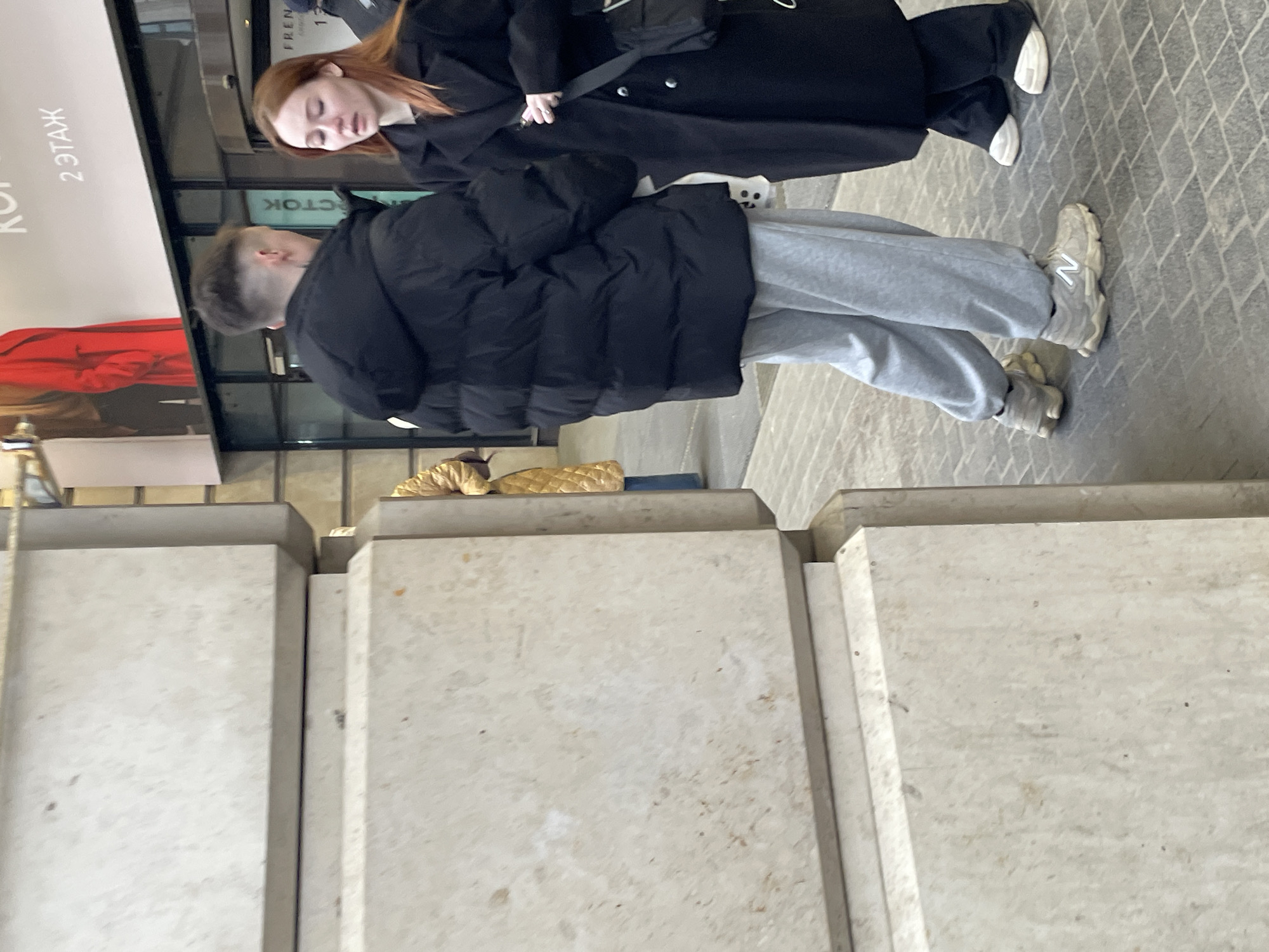The image captures two individuals standing together in what appears to be Saint Petersburg, Russia, based on the Cyrillic text visible on a nearby building. One person, with red hair and wearing a long black coat with a shoulder bag, stands close to the other. The second person is wearing a black puffer jacket and grey sweatpants with New Balance sneakers. They stand on a paved surface, likely a sidewalk, near a building. The foreground contains architectural concrete block elements.