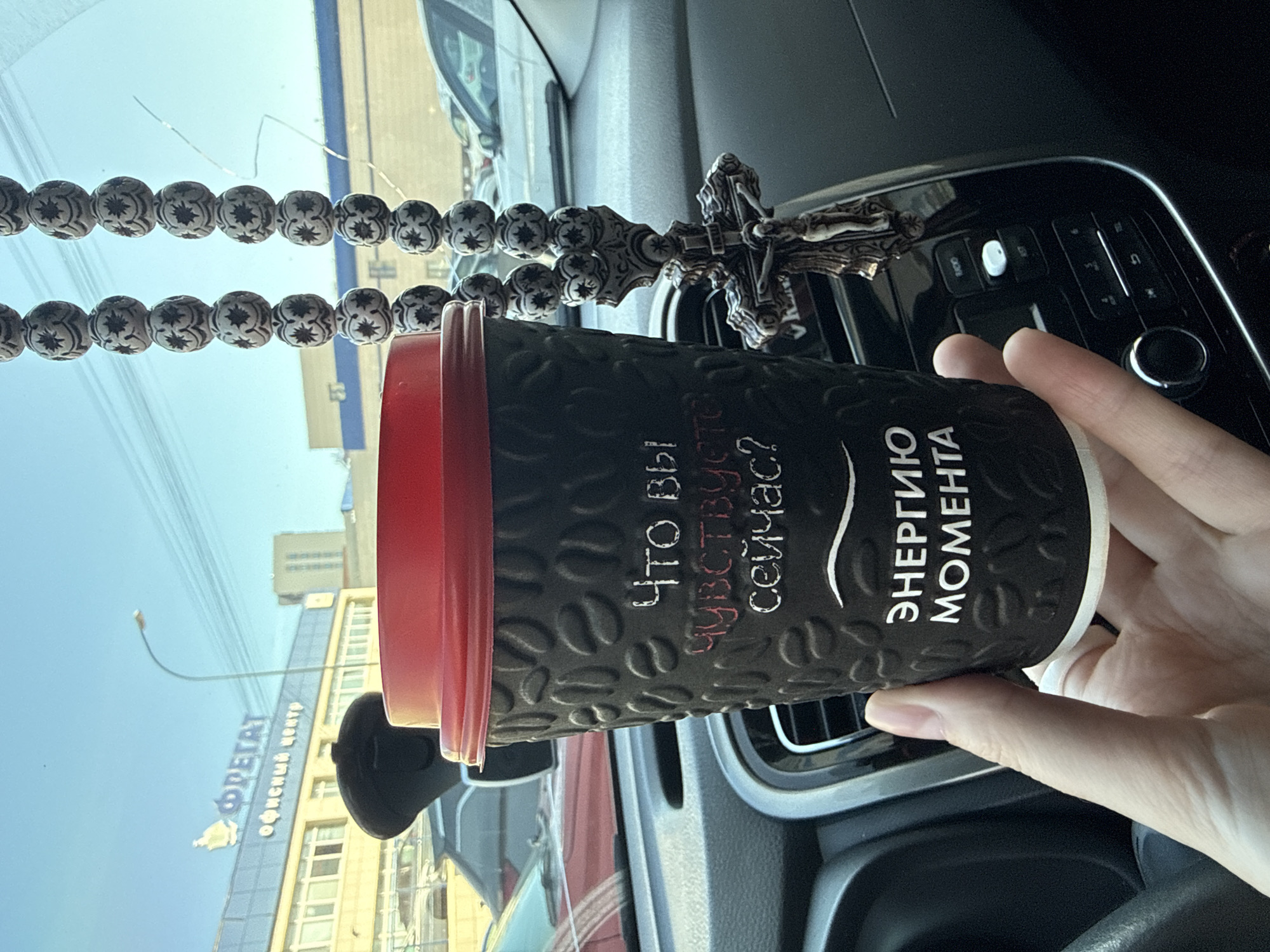 The image shows a person holding a coffee cup in what appears to be the interior of a car. The cup is black with a red lid and has Russian text on it. A string of prayer beads with a crucifix is visible in the background, hanging from the rearview mirror area. Through the windshield, one can see a building with the text "ФРЕГАТ ОФИСНЫЙ ЦЕНТР" (Fregat Office Center), a business center in Izhevsk, Russia. The environment suggests the photo was taken during daylight inside a parked car in Izhevsk, Russia.