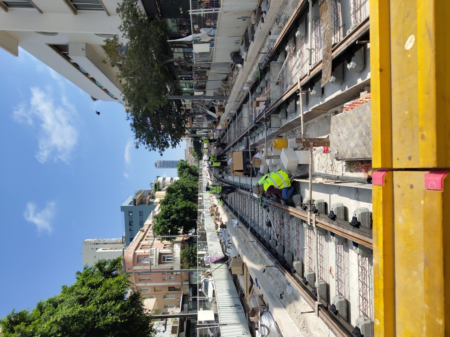 This is a medium shot, eye-level, exterior, daytime photograph of light rail track construction in Tel Aviv, Israel. The scene is set on a street with buildings on both sides, suggesting an urban environment. The foreground is dominated by bright yellow construction barriers, obscuring the very bottom of the frame.
The main focus of the image is a set of parallel light rail tracks being laid down. The tracks are supported by concrete sleepers, and rebar is visible in the unfinished sections, indicating ongoing construction. Several construction workers, all wearing high-visibility yellow vests and hard hats, are actively working on the tracks. One worker in the center is kneeling and appears to be tightening a bolt or making an adjustment to the track. Another worker in a similar vest is standing further down the track. In the background, other workers are visible, as well as piles of construction materials, including concrete blocks and wooden planks.
The street is lined with buildings. On the left, there are multi-story buildings with balconies and trees. On the right, a modern white building with balconies is prominent. The sky is blue with scattered white clouds. The overall impression is one of a busy construction site in an urban setting. The presence of the light rail construction strongly suggests this is an area undergoing significant infrastructure development.