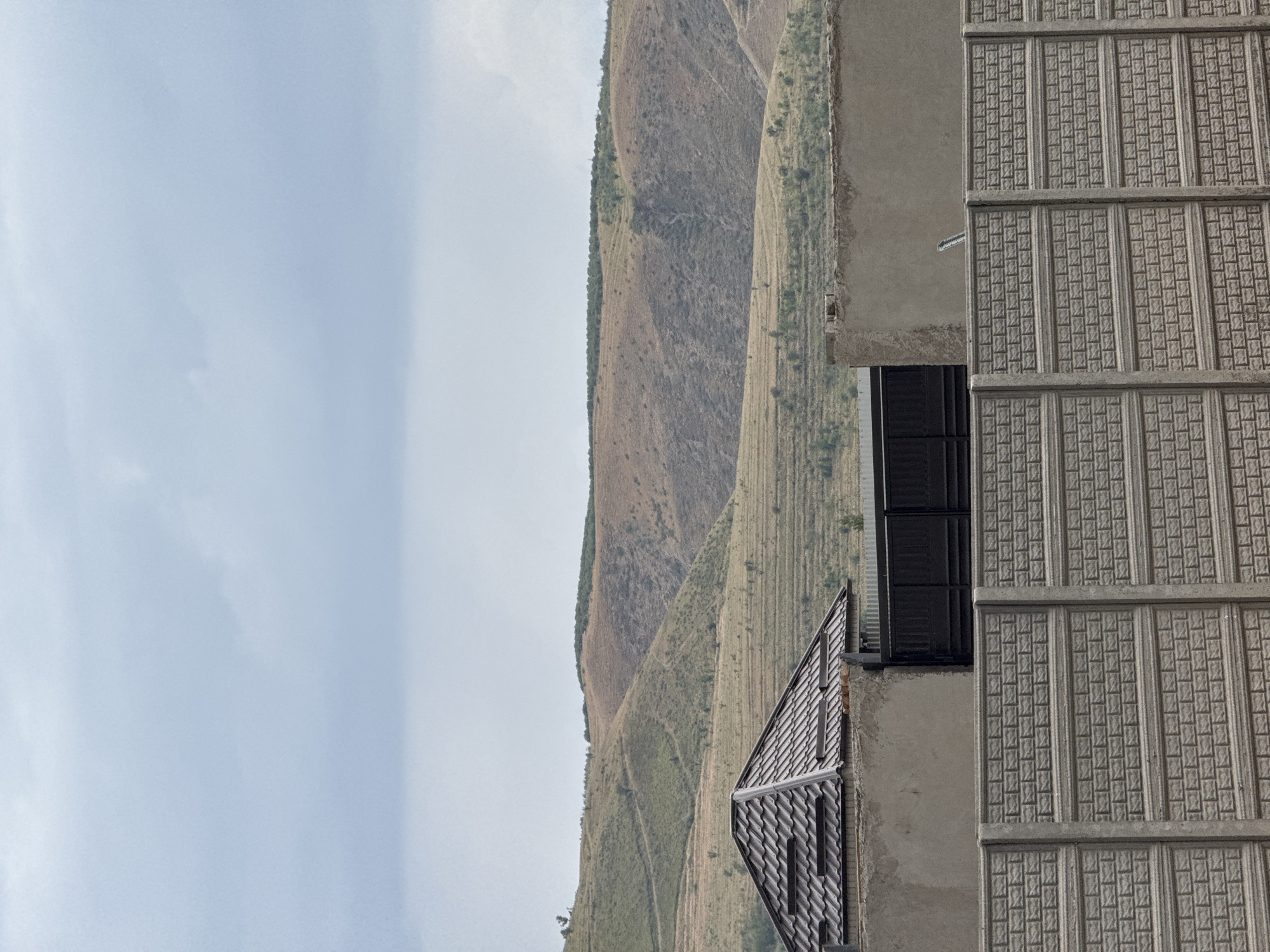 This media file shows a view of a building's facade and a mountain landscape. The building has a textured, gray concrete exterior with decorative panels and a window with dark shutters. A section of the building's roof, made of dark, overlapping tiles, is also visible, with a metal railing in front of it. Beyond the building, the scene transitions to a vast mountain range covered in dry vegetation, with terraced slopes suggesting agricultural activity or erosion patterns. The sky is overcast, with a soft, diffused light indicating daytime, possibly late afternoon or early morning, with no clear shadows. The overall impression is one of a remote or mountainous region, with a contrast between man-made structures and the natural environment. No people or specific activities are depicted. There is no visible text in the image.