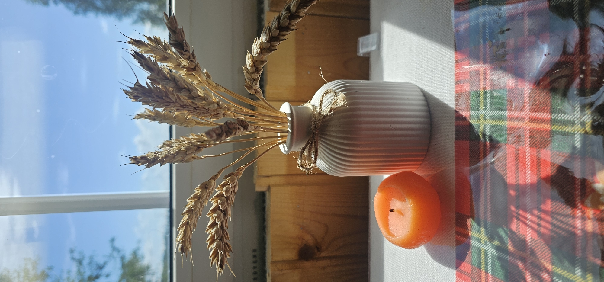 An orange, spherical candle sits to the left of a ribbed white ceramic vase filled with dried wheat stalks. The vase is tied with twine. The objects are placed on a white tablecloth with a red, green, and yellow plaid pattern overlaying it. The scene is illuminated by sunlight streaming through a window, casting shadows. In the background, wooden slats are visible, and beyond that, a bright blue sky with some clouds can be seen through the window. The location is the city of Tikhonovo SNT, Russia.