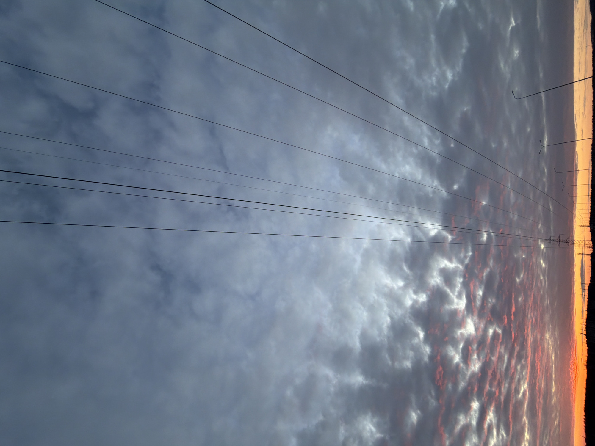 An outdoor scene captured in Chaikovsky, Russia, features a sky dominated by a combination of altocumulus and stratocumulus clouds during either sunrise or sunset. Approximately ten to twelve dark, thin electrical transmission lines extend diagonally from the upper-left to the lower-right of the frame. The upper portion of the sky exhibits muted grey-blue clouds, while the lower portion, near the horizon, is illuminated by vibrant orange and red light, indicating the sun's position below the visible frame. The horizon presents a dark silhouette of the landscape, featuring a prominent metal lattice electrical transmission tower on the left side. To the right of the tower, a series of at least five streetlights with slender, curved poles are visible in silhouette, suggesting a road or developed area. No individuals or specific activities are present in the image.