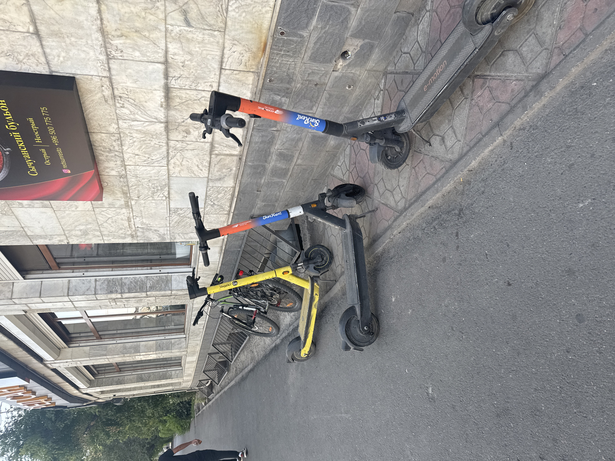 In Bishkek, Kyrgyzstan, a street scene unfolds with a focus on personal transportation. Three electric scooters are parked along a tiled sidewalk next to a building with a stone facade. The scooters, two of which are branded "SunRent," exhibit colorful designs. One scooter is black with orange and blue accents, while another features a gradient of blue and orange. The third scooter, yellow, stands out among them, with a bicycle leaning against it. A person in dark clothing is walking away from the camera on the asphalt road, adding a human element to the urban environment. The building's exterior is characterized by stone tiles and large windows, suggesting a commercial or public space.