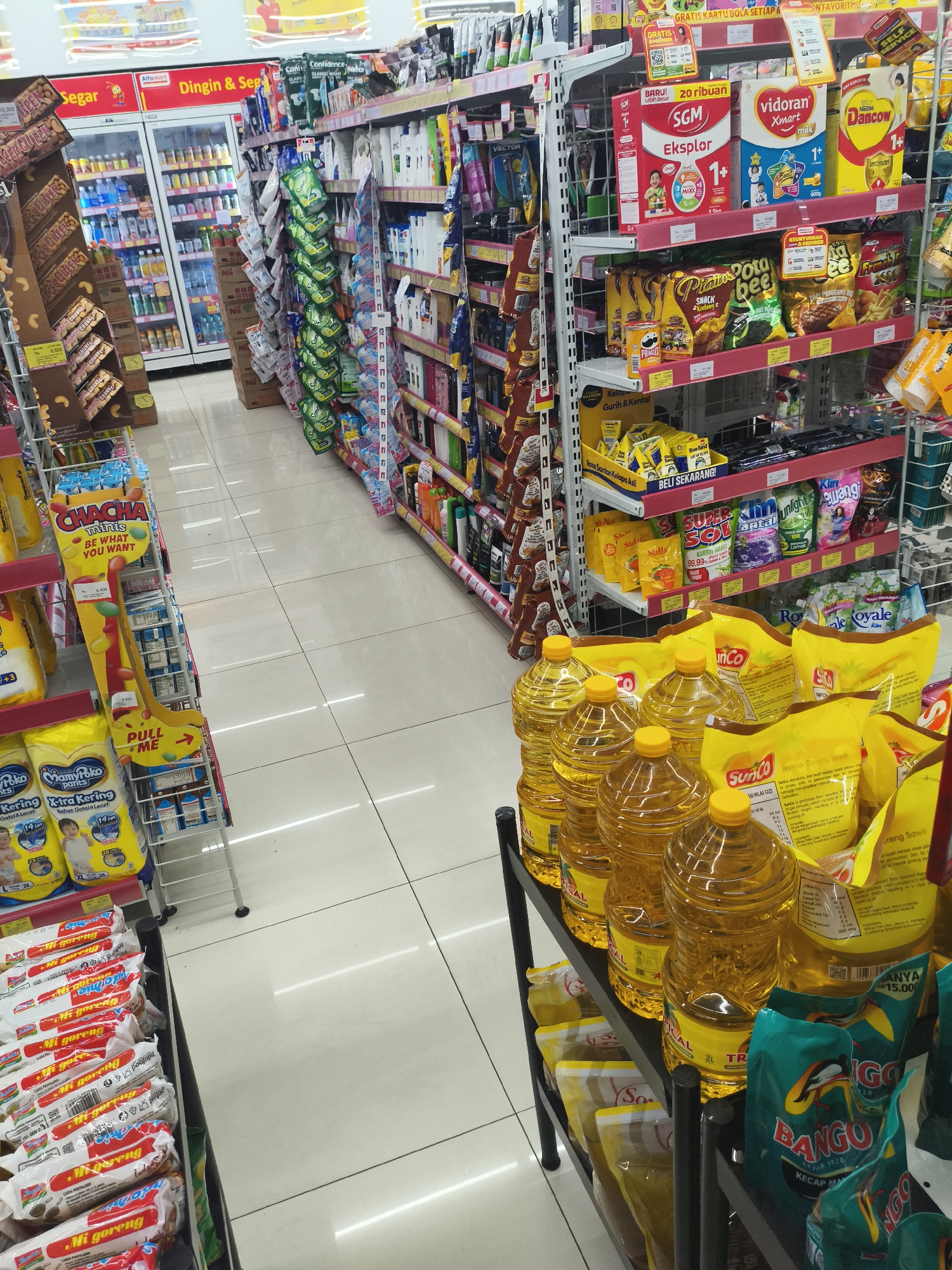 The image shows an interior view of a brightly lit convenience store in Depok, Indonesia. The perspective is from a low angle, looking down an aisle lined with shelves stocked with various consumer goods. On the left, refrigerators contain beverages, and shelves display chocolate bars and other packaged snacks. The center aisle is filled with a diverse array of products, including infant formula, snacks, cleaning supplies, and cooking oil. The flooring is white tile. The overall scene depicts a typical retail environment with merchandise neatly arranged on display shelves.